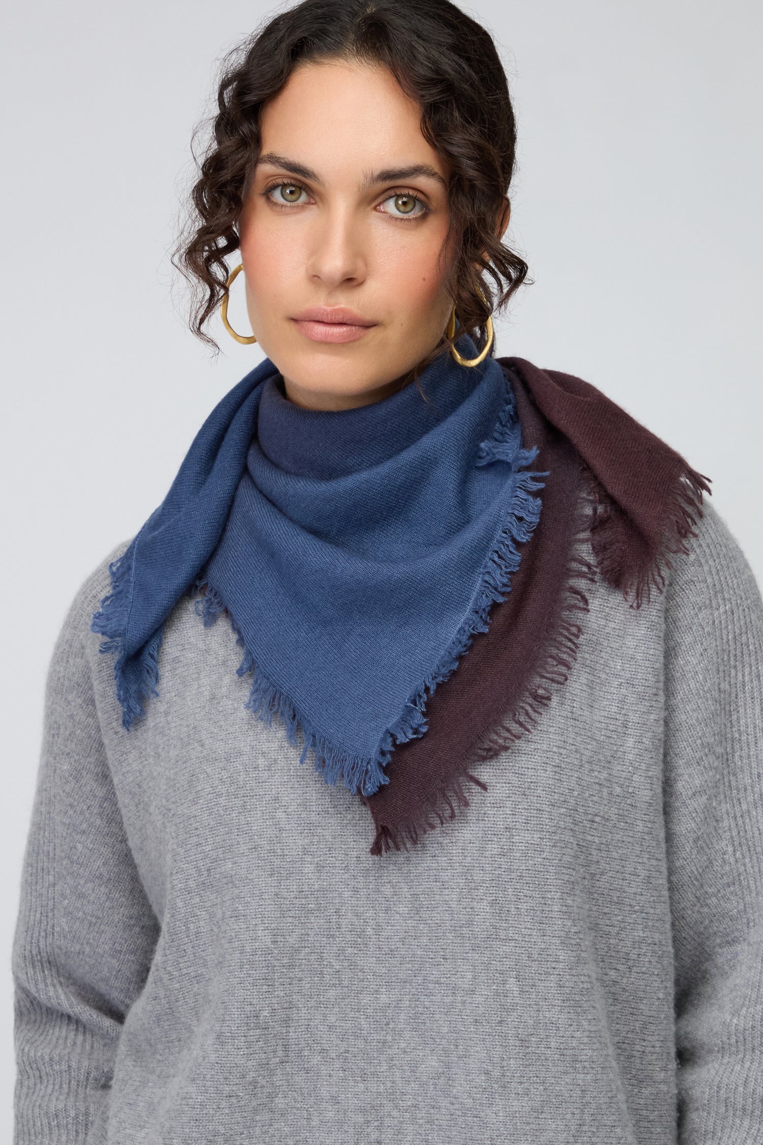 A woman in a grey sweater wears the Ombre Cashmere Square Scarf in blue and brown tones with gold hoop earrings, standing against a plain background.