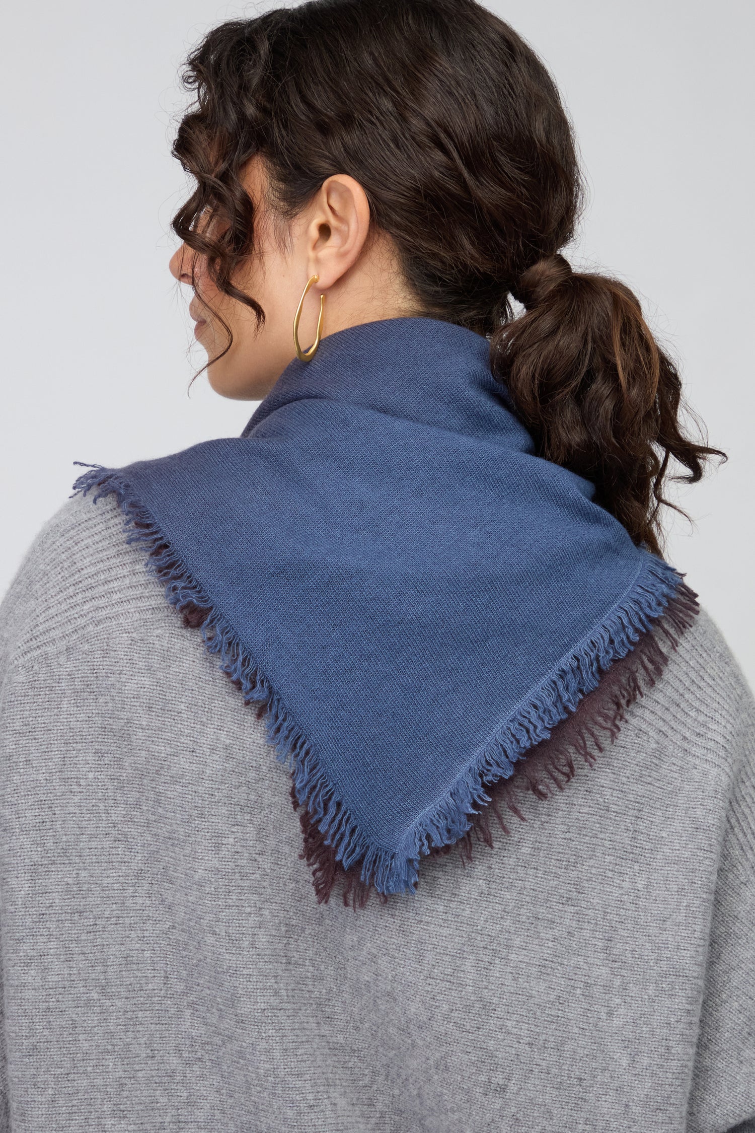 Woman with curly brown hair in a low ponytail, wearing gold hoop earrings, a grey sweater, and the Ombre Cashmere Square Scarf—luxurious blue, square-shaped with fringe—photographed from behind.