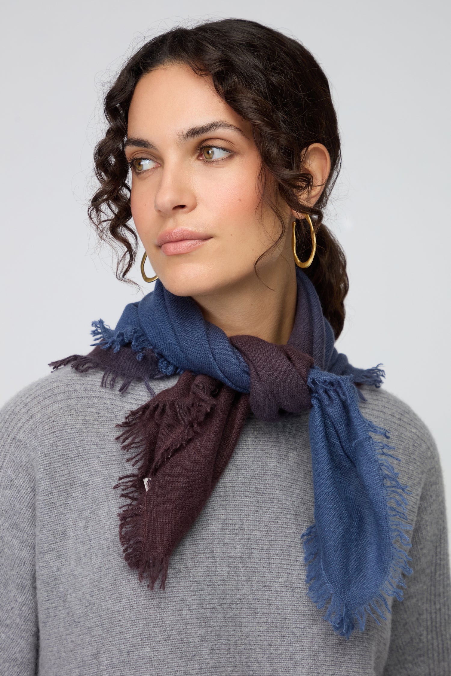 A woman with wavy brown hair, gold hoop earrings, and a grey sweater wears the Ombre Cashmere Square Scarf in blue and brown fringe, looking slightly to the side.