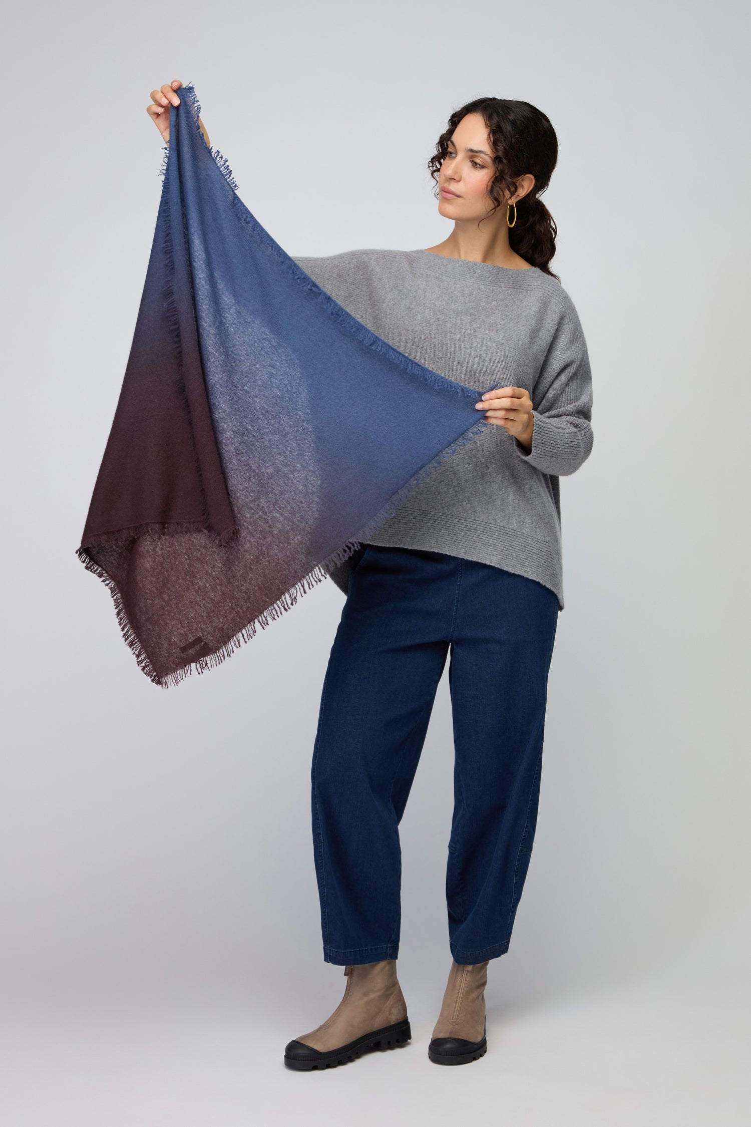 A person in a gray sweater and blue jeans displays the Ombre Cashmere Square Scarf, featuring blue and burgundy hues with fringed edges—a luxurious, stylish addition to any wardrobe.