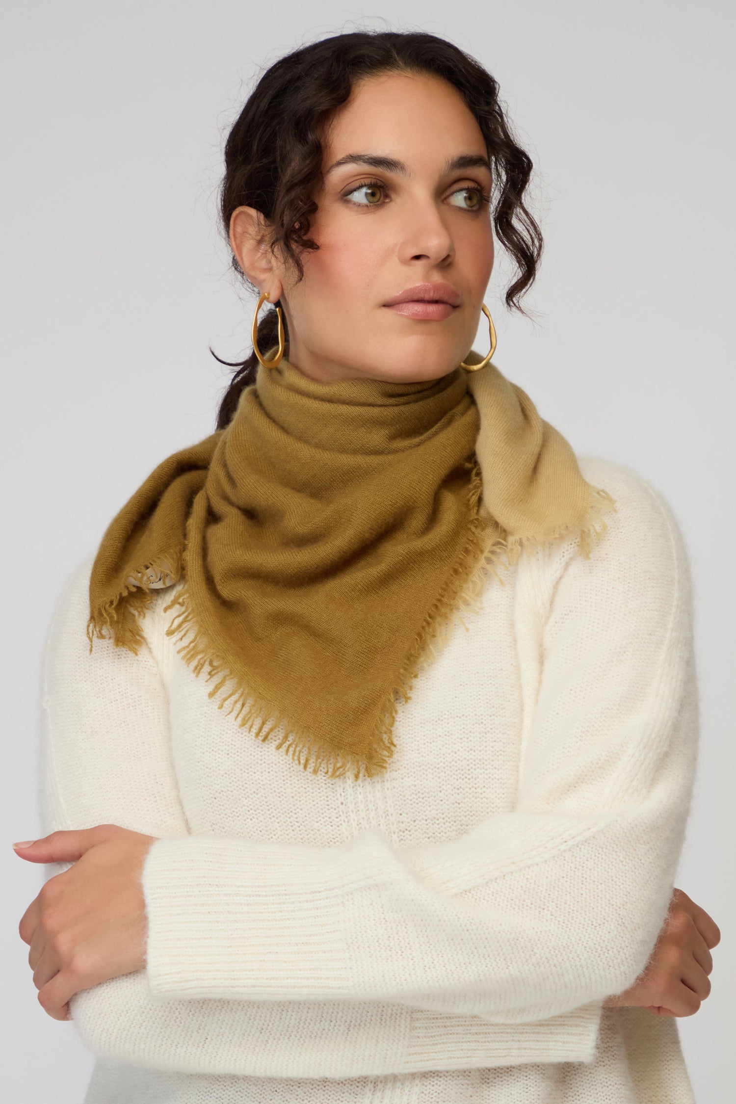 A woman wearing a white sweater and an Ombre Cashmere Square Scarf in tan looks to her left with her arms crossed.