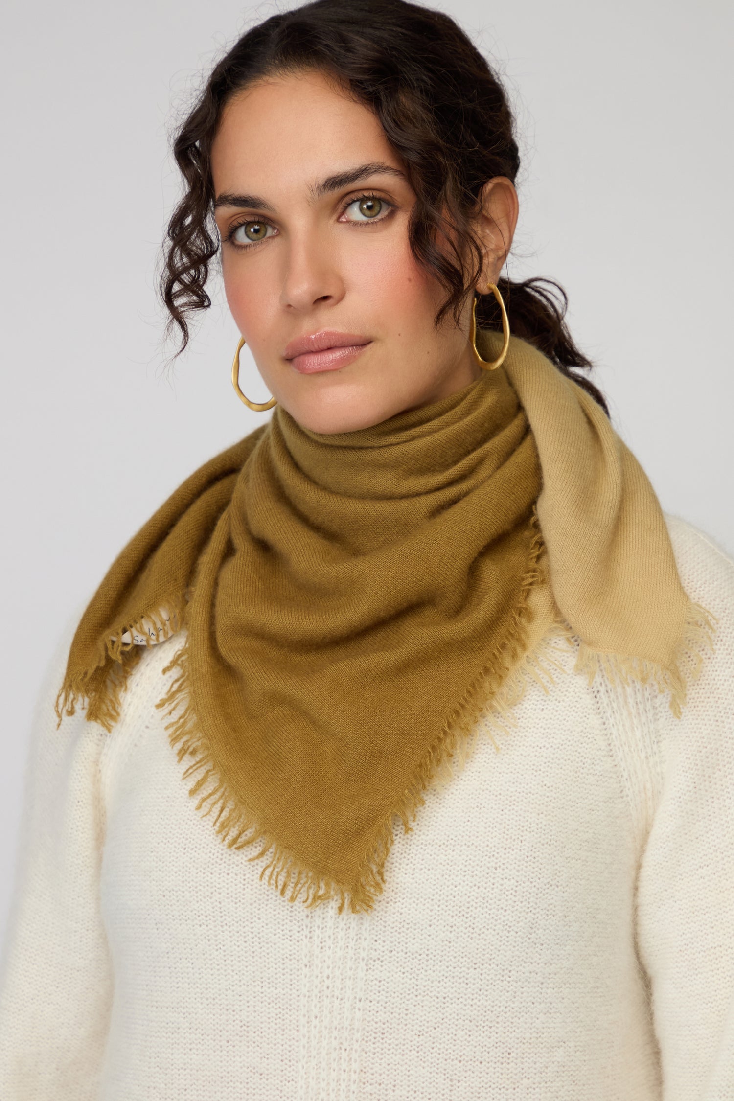 Woman wearing a white sweater and the Ombre Cashmere Square Scarf with fringe, paired with gold hoop earrings, looking at the camera against a plain background.