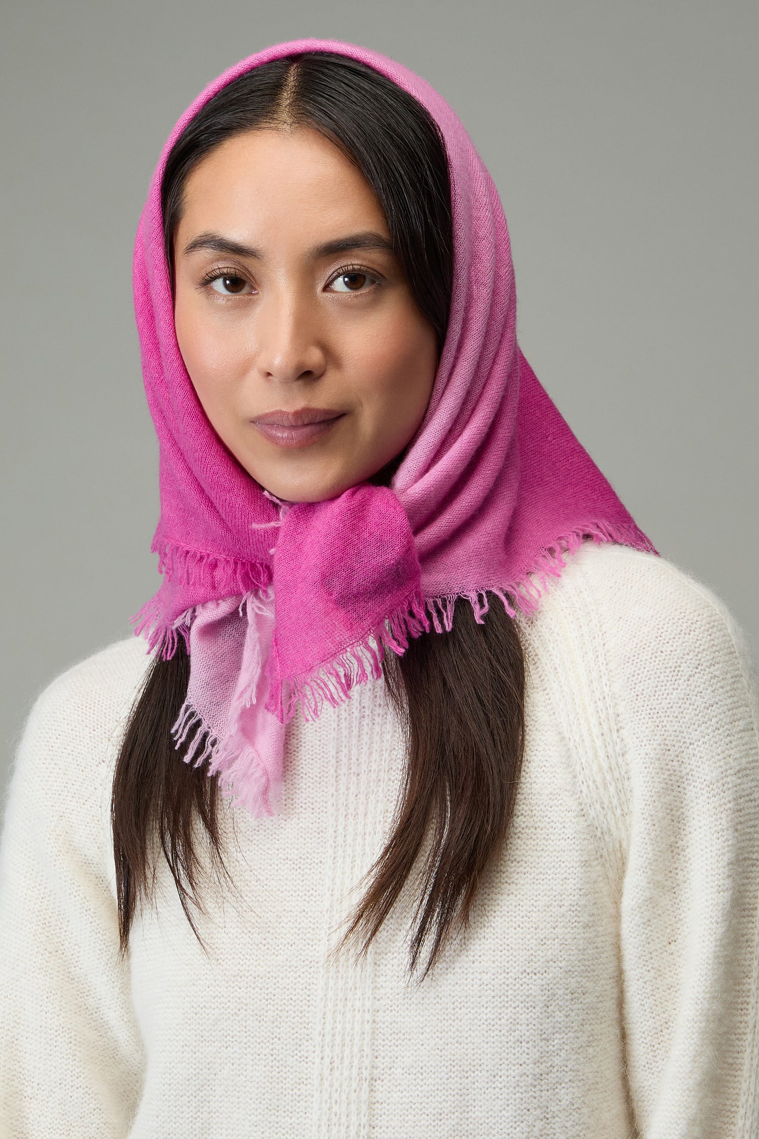 Woman wearing the Ombre Cashmere Square Scarf in pink with a white sweater, looking at the camera against a plain gray background.