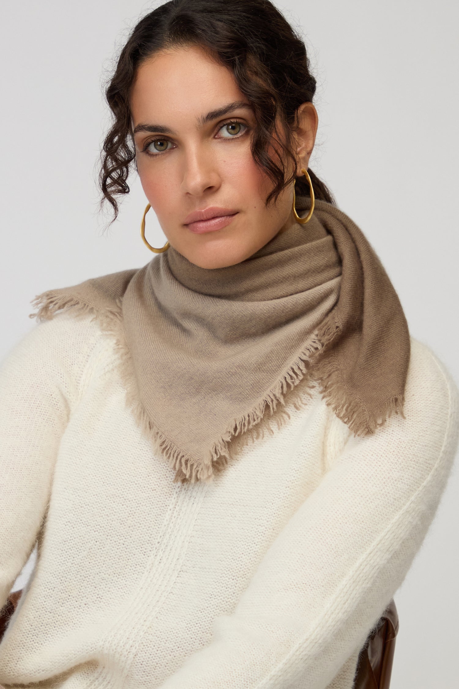 A woman with wavy brown hair, gold hoop earrings, and a cream sweater wears the Ombre Cashmere Square Scarf while sitting against a plain light background.