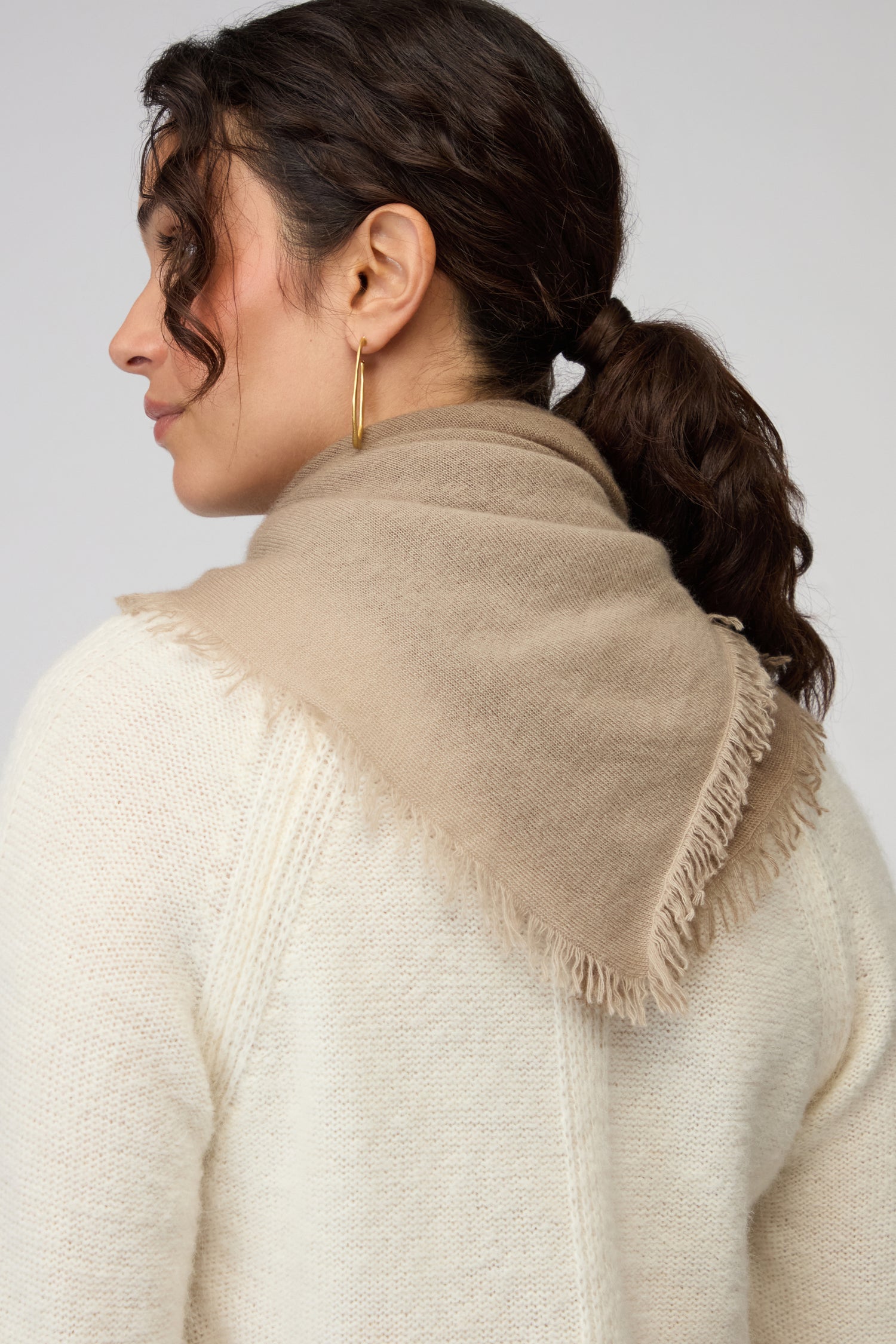 A woman with dark hair in a ponytail is shown from the back and side, wearing gold hoop earrings, a cream knit sweater, and the Ombre Cashmere Square Scarf with beige fringe.