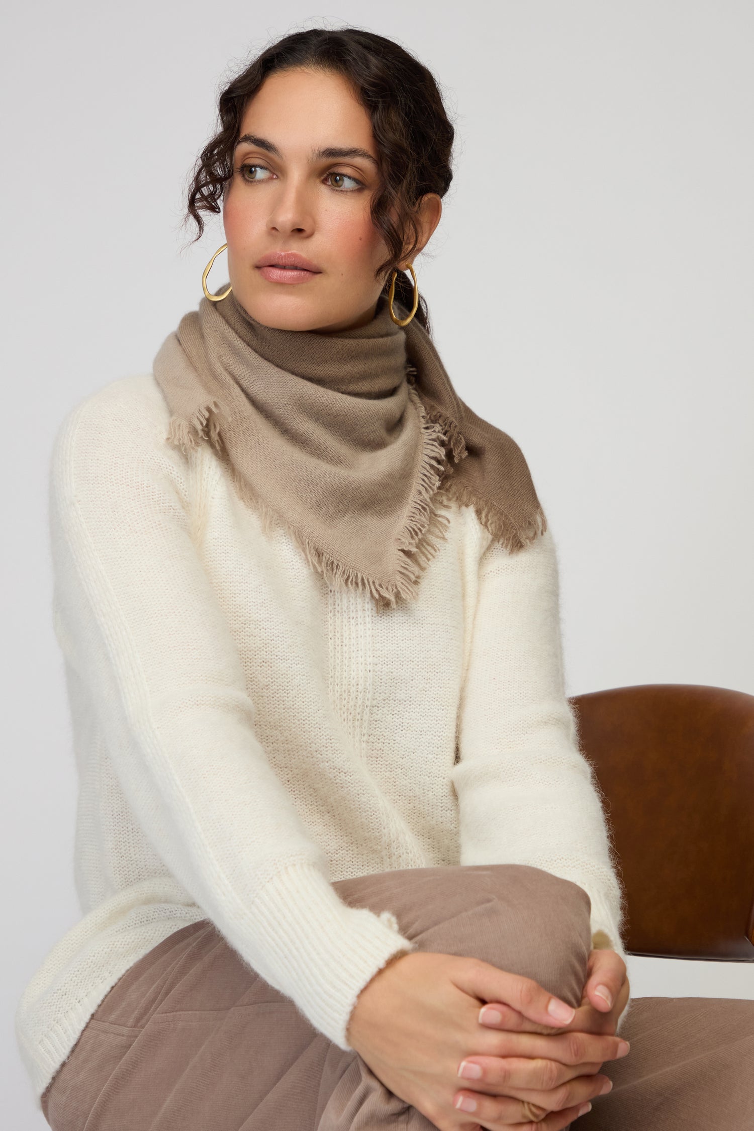 A woman in a cream sweater and an Ombre Cashmere Square Scarf sits on a brown chair, her arms resting on her knee as she gazes to the side.