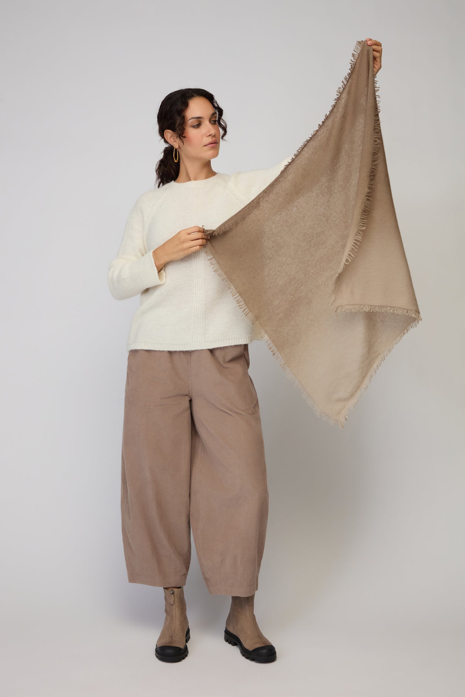 A woman holds up the Ombre Cashmere Square Scarf, a large beige luxury piece with fringe. She pairs it with a cream sweater, loose tan pants, and brown ankle boots against a simple backdrop.