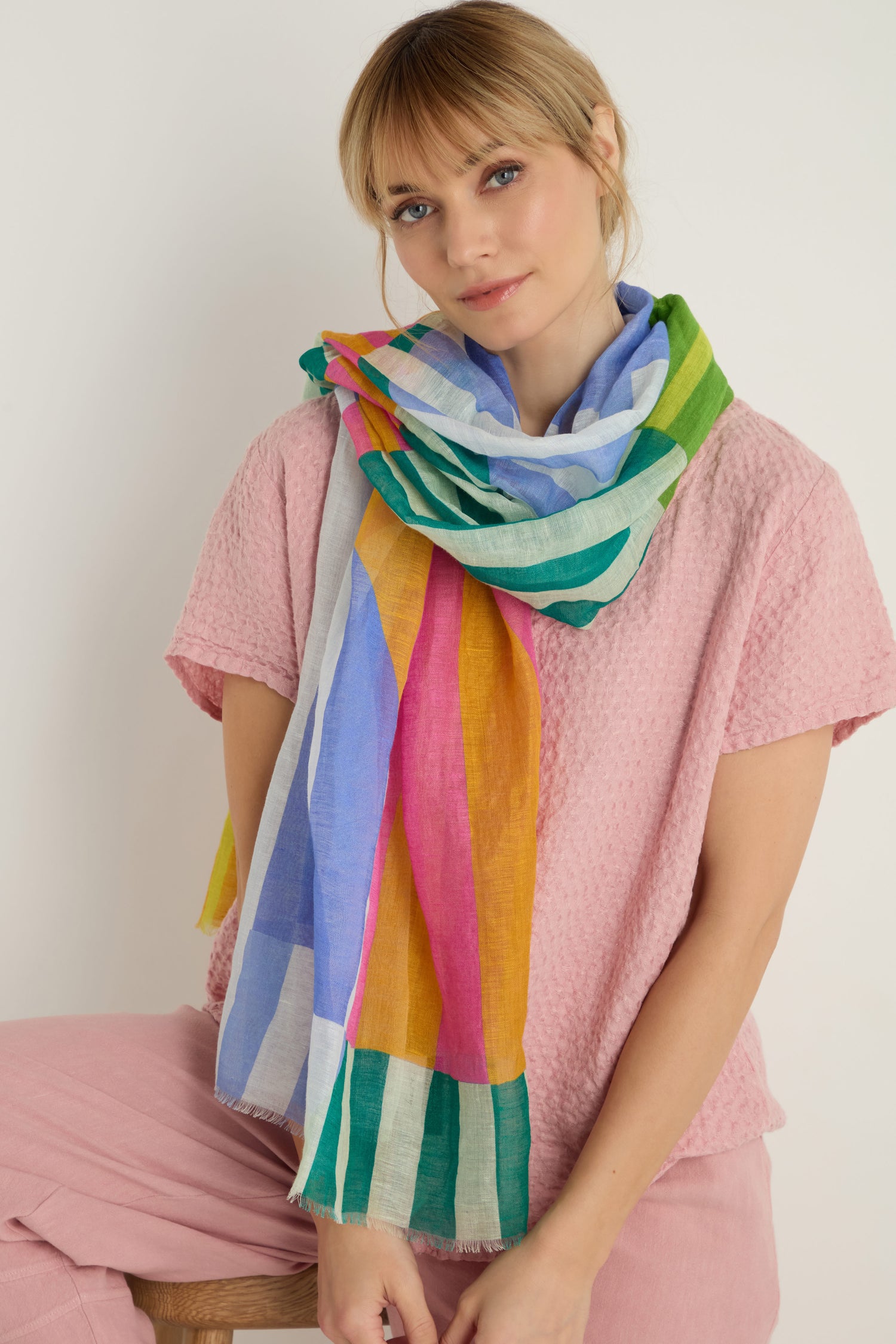 A woman in a textured pink top and pants sits on a stool, modeling the Sunburst Block Print Linen Blend Scarf wrapped loosely around her neck.