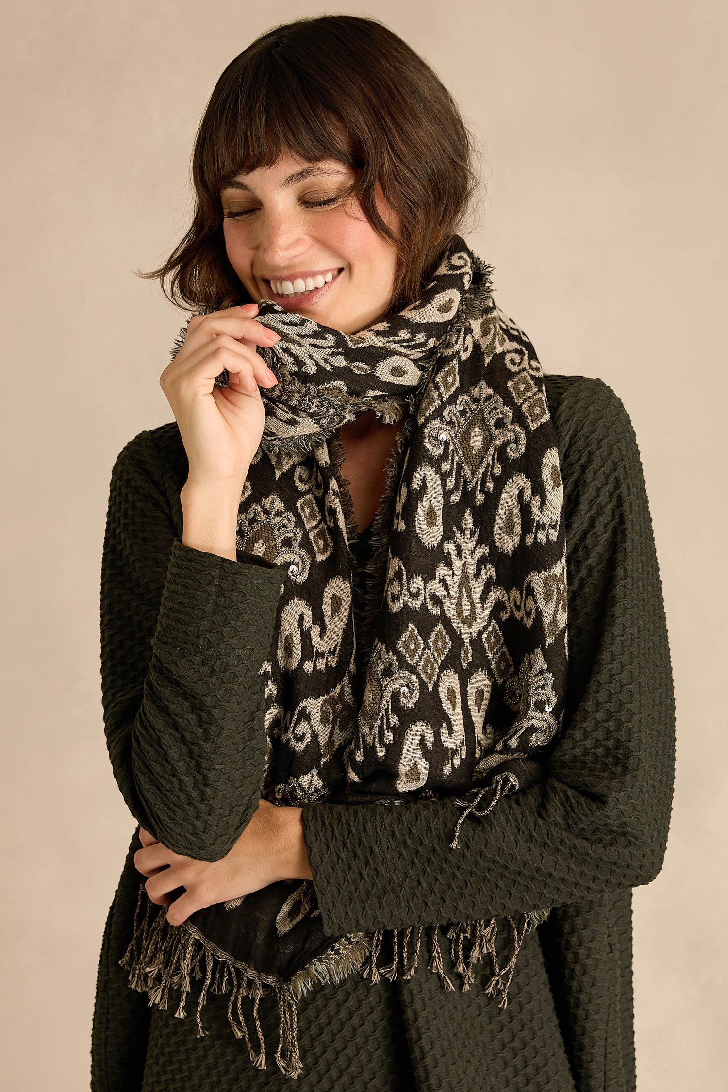 A woman smiles with her eyes closed, standing against a neutral palette background, wearing the Motif Sequin Scarf styled with a textured dark green cotton-wool blend sweater.