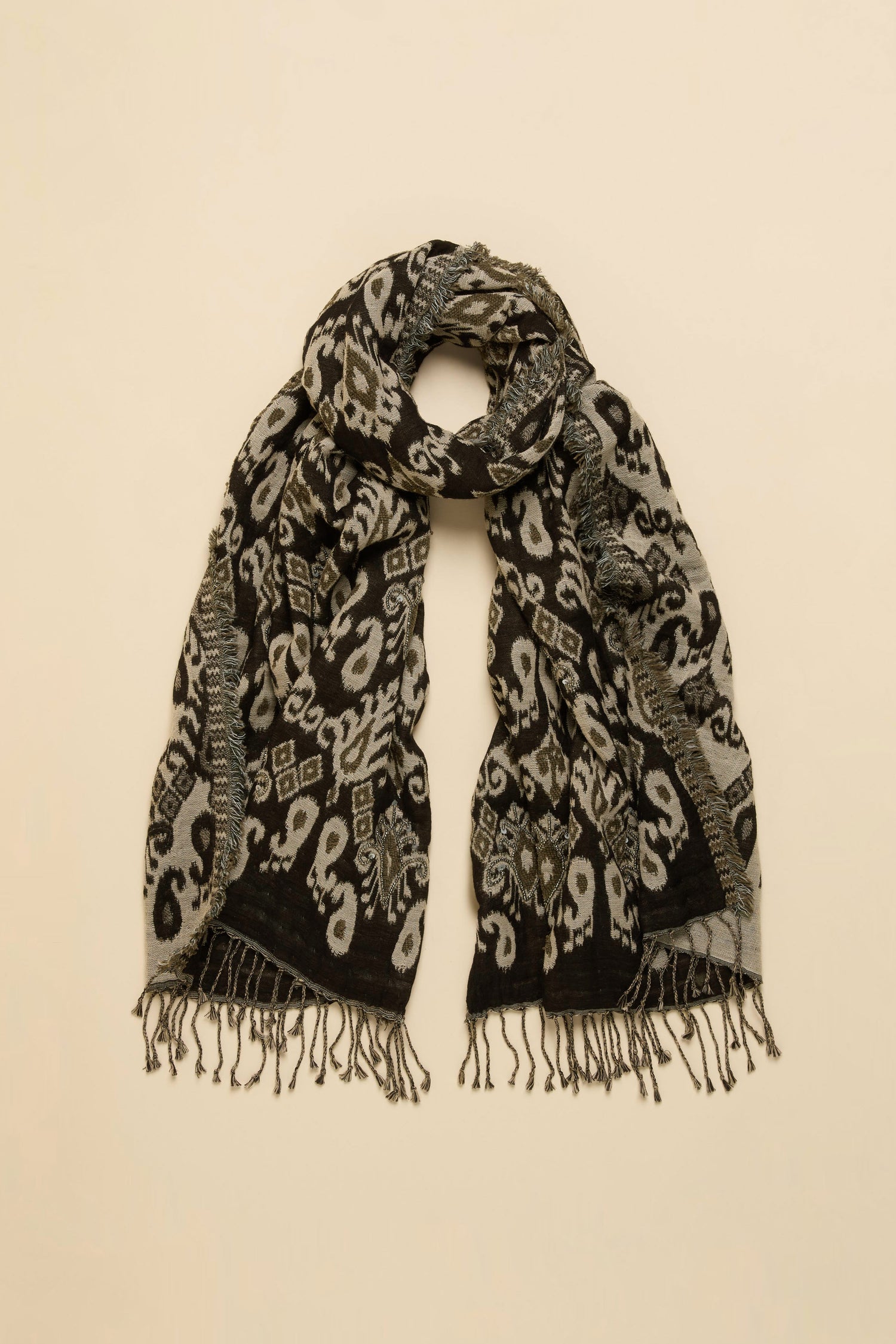 The Motif Sequin Scarf, featuring a black and gray pattern with fringed ends and crafted from a soft cotton-wool blend, is laid flat on a beige background.