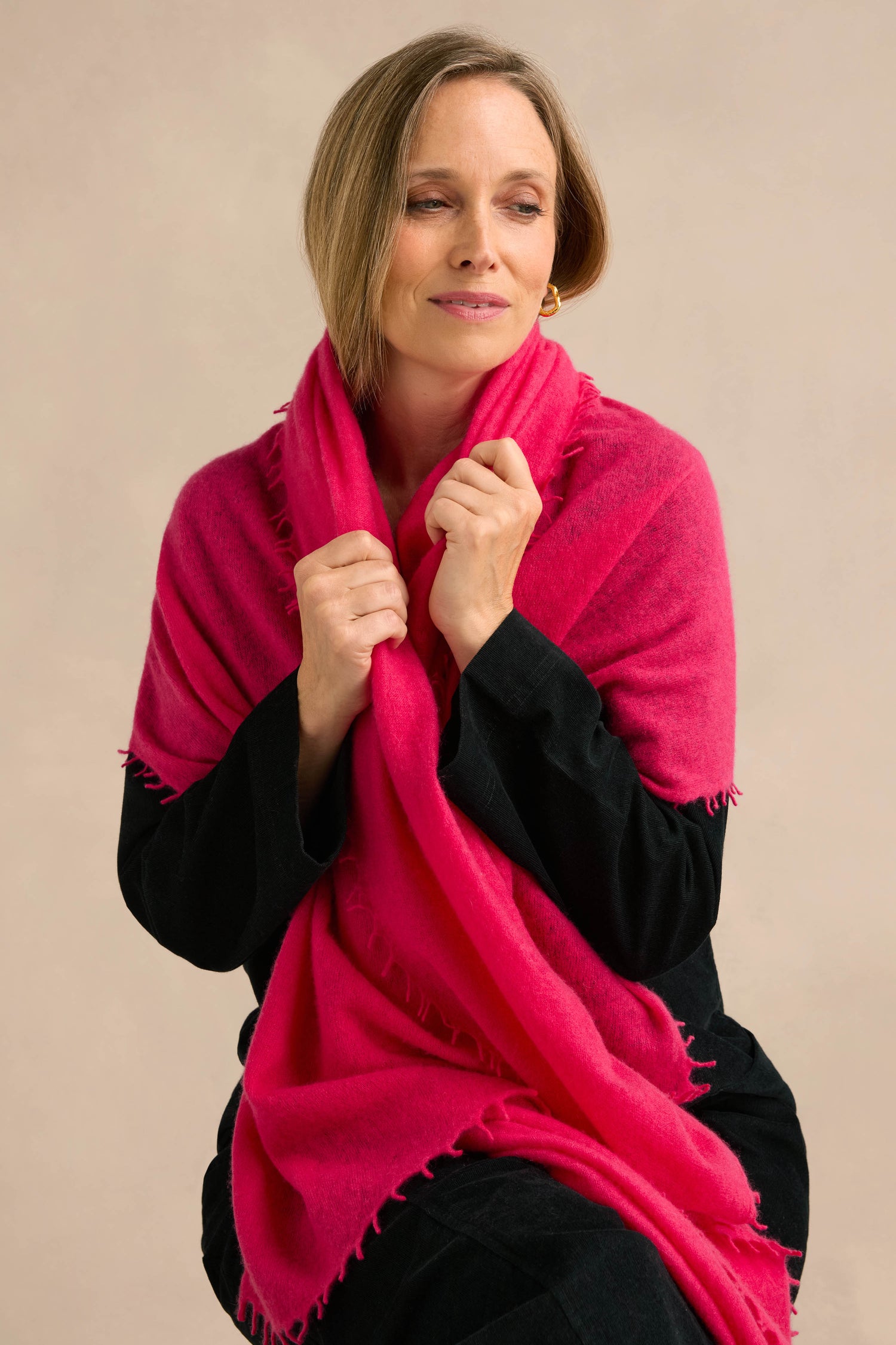 A woman with short hair wears the Fuchsia Soft Cashmere Scarf over a black outfit, holding the luxurious accessory close with both hands. She gazes slightly downward and to the side against a neutral background.