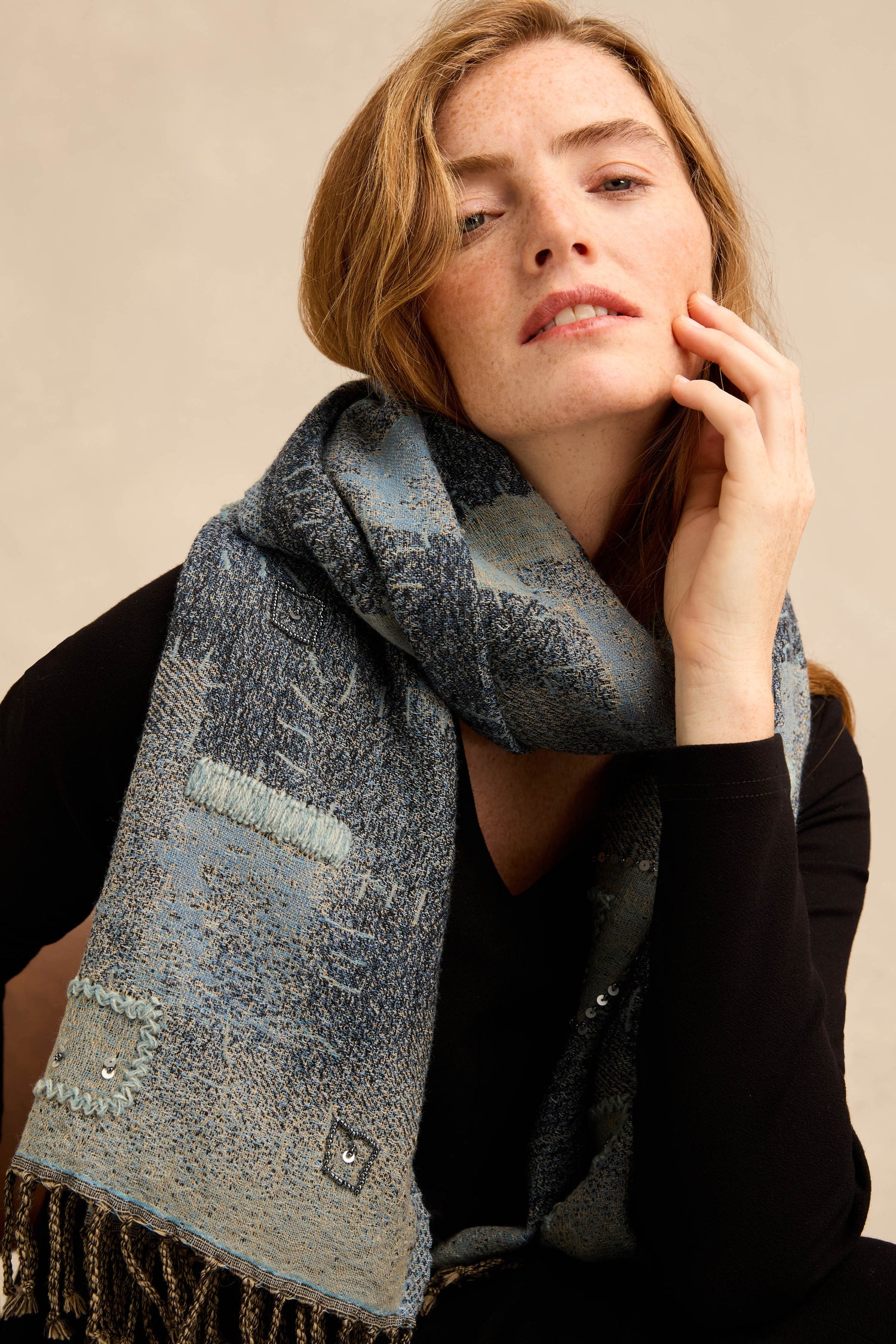 A woman in a black top wears the Etched Patchwork Sequin Wool Scarf—a textured blue wool scarf featuring stitched details, subtle patchwork design, and fringe—as she poses with her hand gently touching her face.