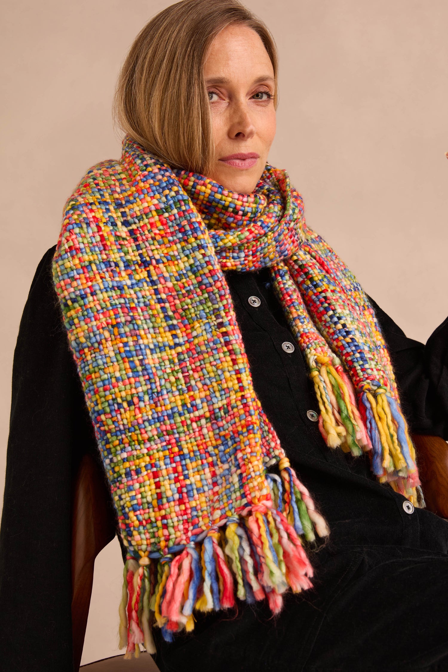 A woman is wearing a black button-up top and the Rainbow Tassel Scarf, crafted from soft brushed yarn, seated against a neutral background. This stylish accessory offers lightweight warmth while adding a vibrant touch to her ensemble.