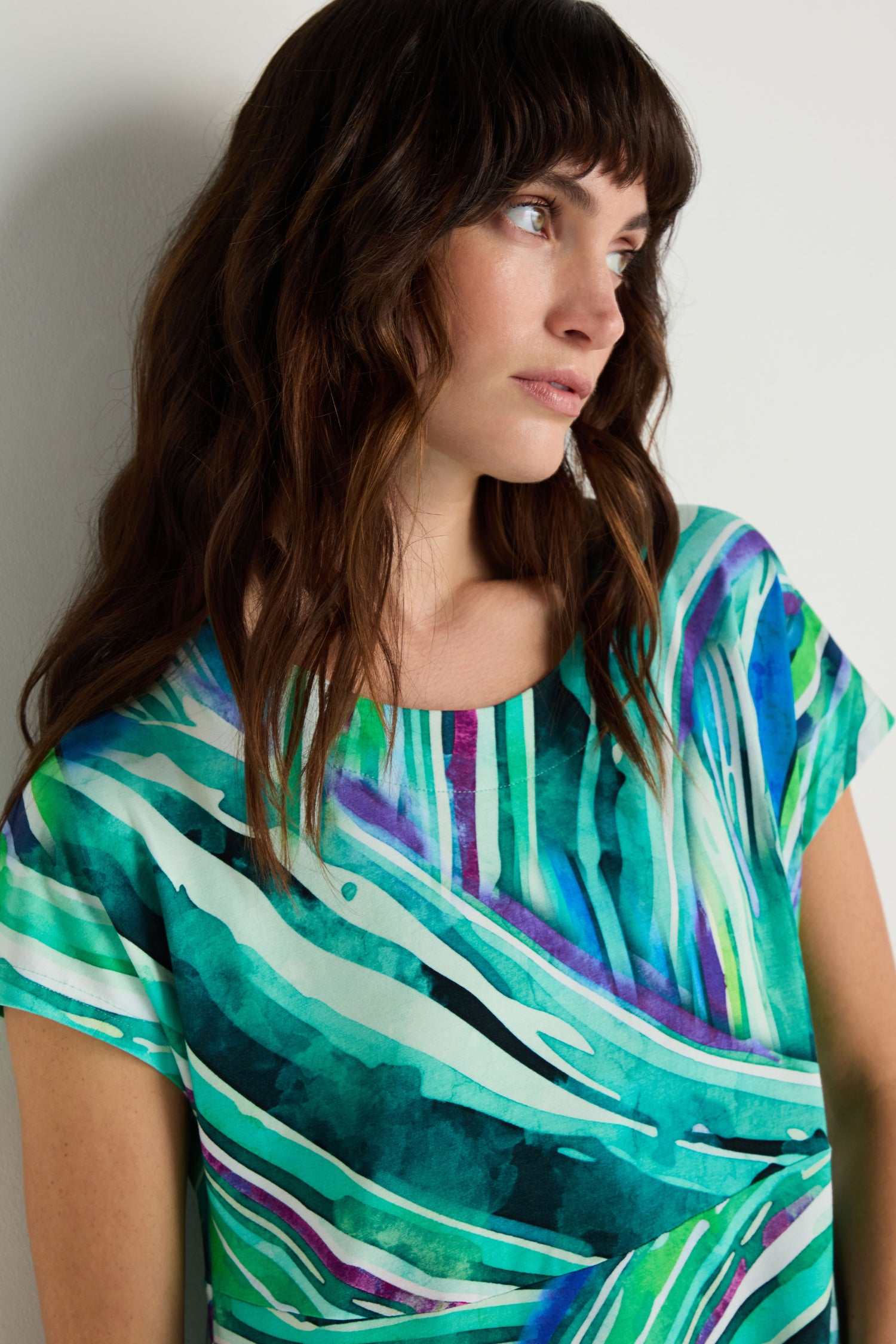 Woman with wavy brown hair looks to the side, wearing the Blurred Lines Swing Dress—a short-sleeved, stretch jersey dress featuring green, blue, and white abstract patterns.