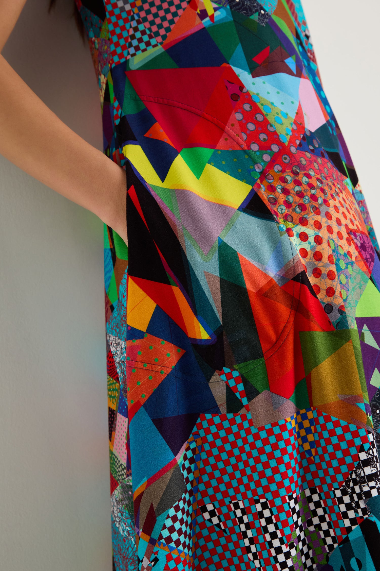 A person wears the Quilt Print Flared Dress—a colorful, geometric-patterned stretch jersey design—stands against a light wall with one hand in a pocket.