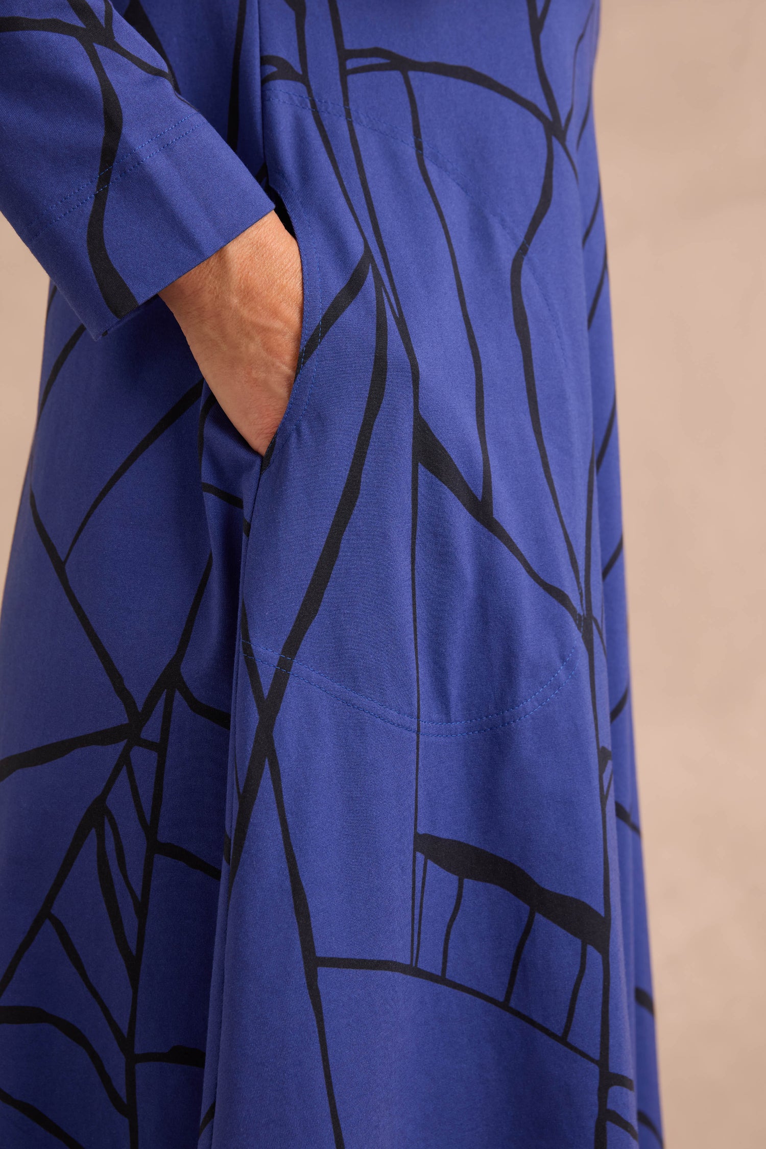 A person’s hand resting in the side pocket of the Linear Print Jersey Flared Dress Aino, a blue dress featuring a black abstract line pattern, photographed against a neutral background.