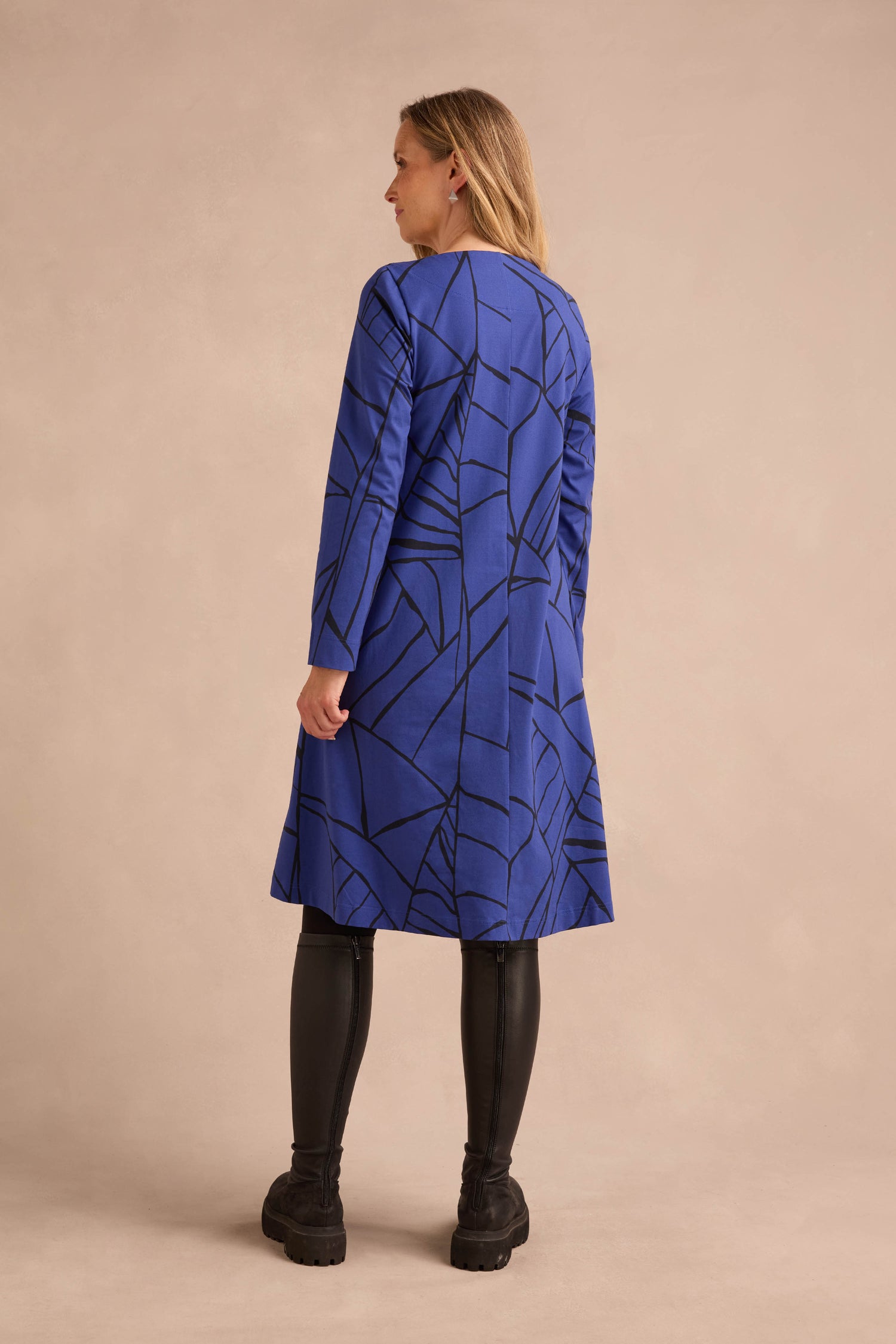 A woman stands facing away, wearing the Linear Print Jersey Flared Dress Aino in blue with a black abstract pattern, paired with black leggings and black shoes, against a beige background.
