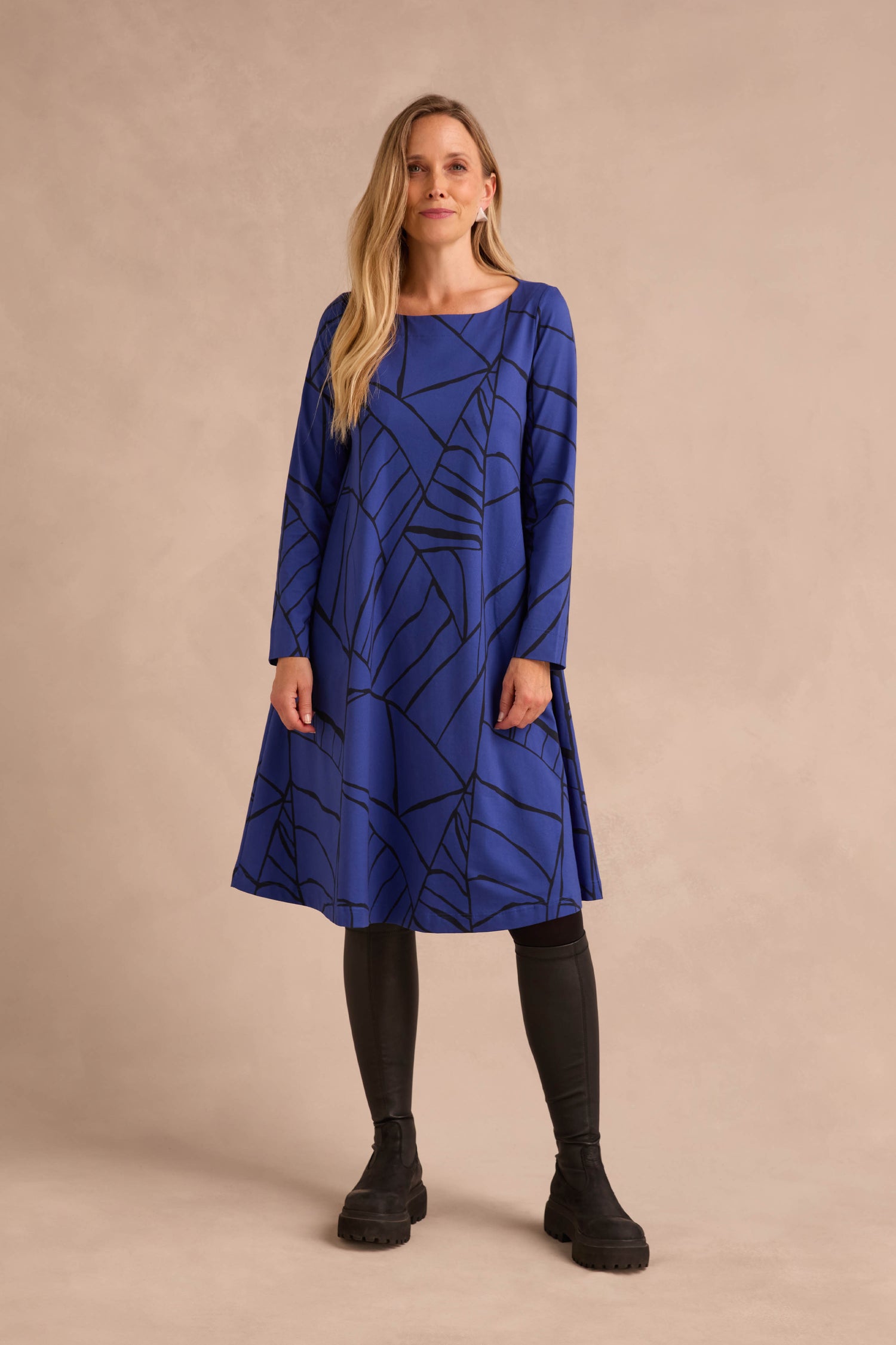 A woman stands against a neutral beige background wearing the Linear Print Jersey Flared Dress Aino, featuring black geometric lines, styled with black leggings and black boots.