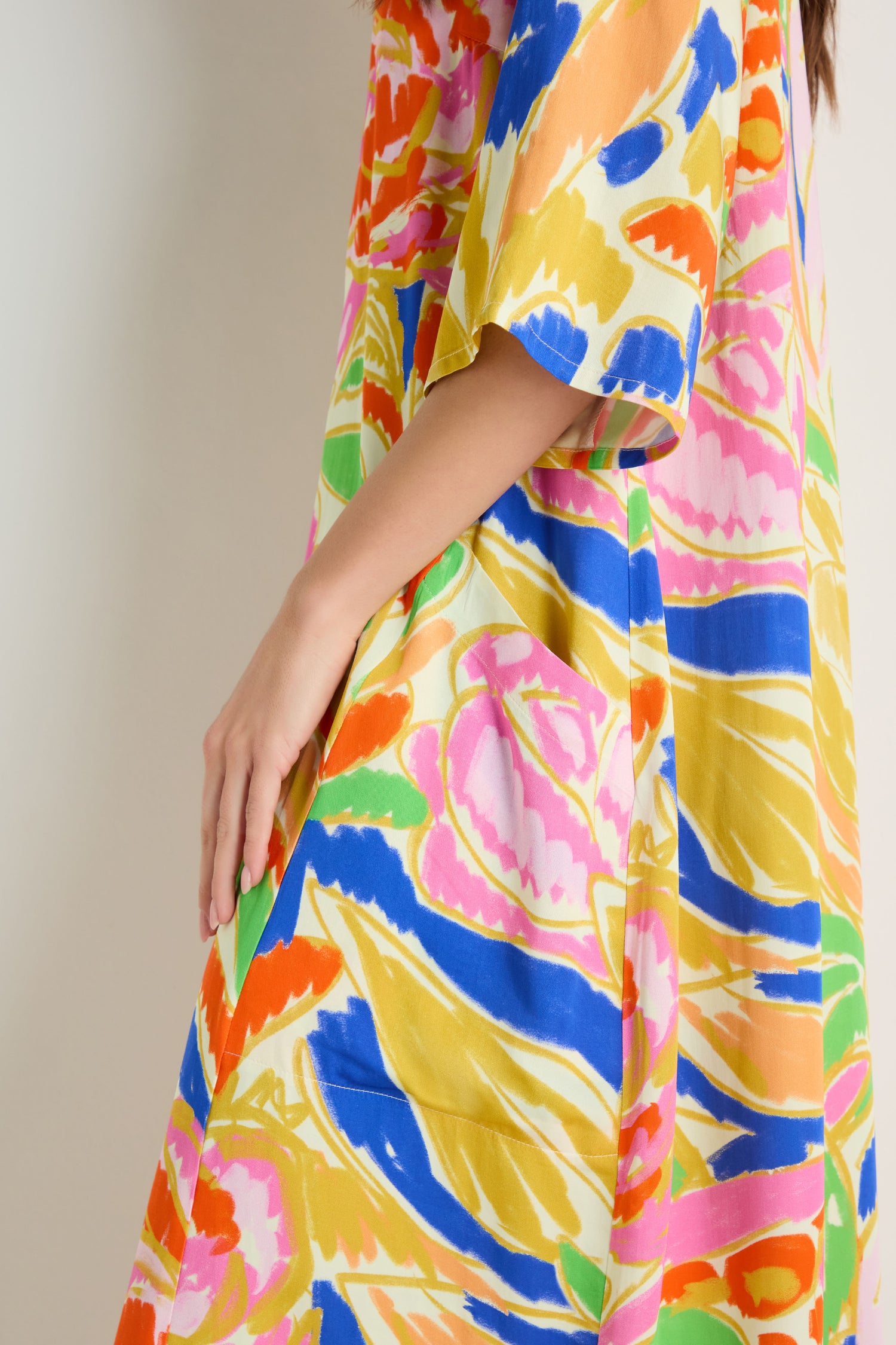 A person wearing the Abstract Floral Pleated Dress, a woven viscose midi with an abstract floral print in blue, pink, orange, yellow, and green. Only their torso and arm are visible against a neutral background.