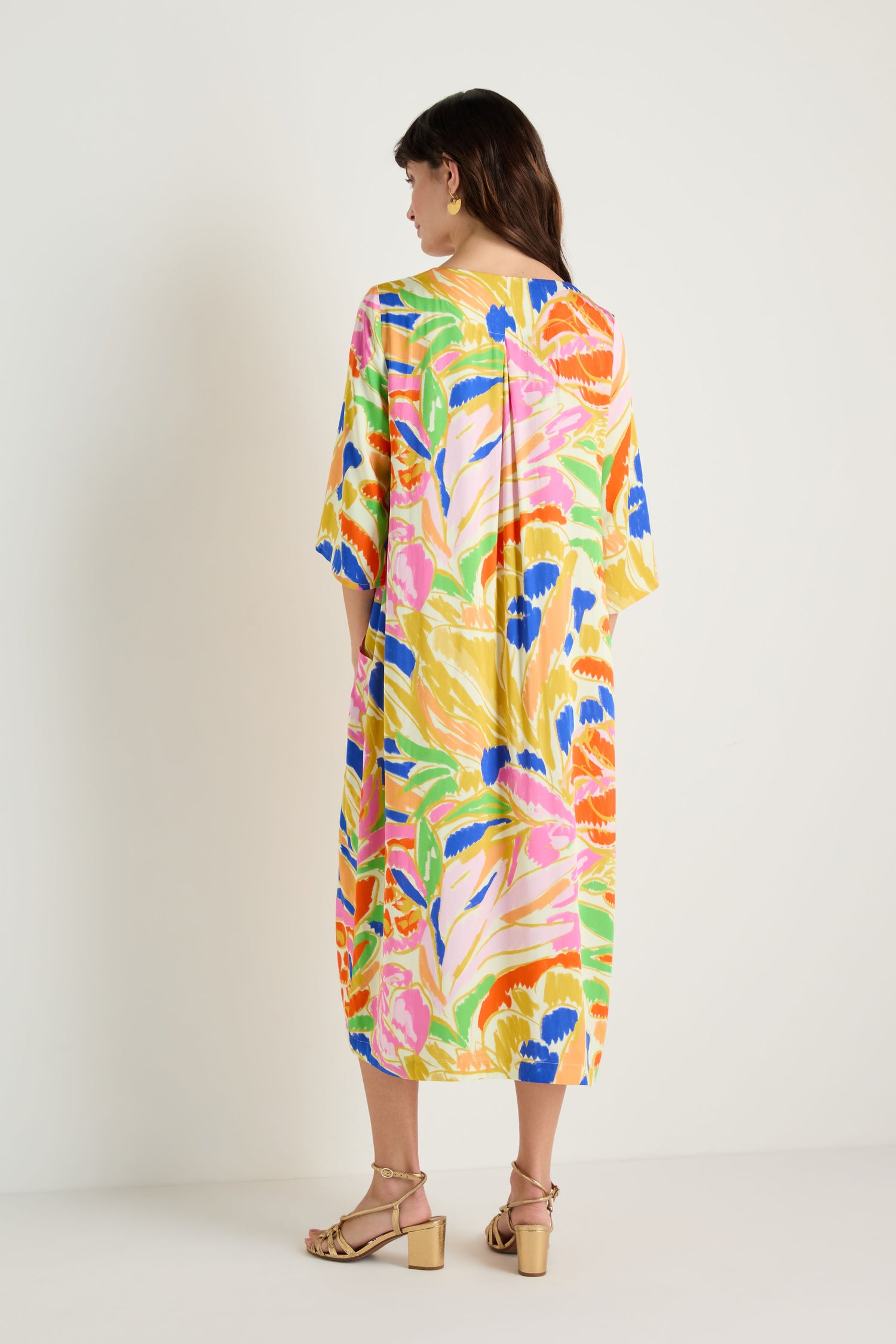 A woman faces away in an Abstract Floral Pleated Dress with elbow-length sleeves and beige heeled sandals, set against a plain white background.