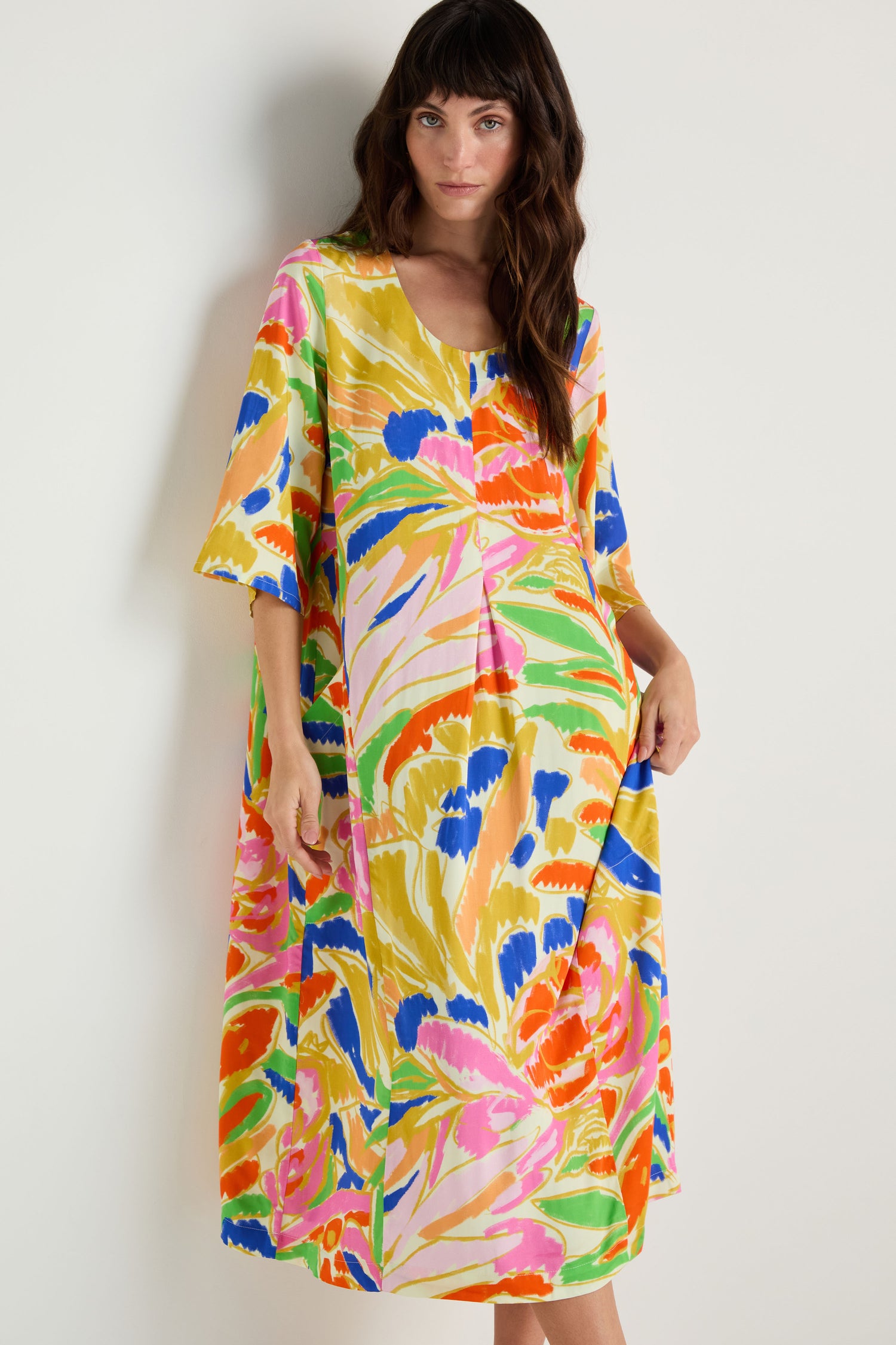 A woman models the Abstract Floral Pleated Dress, a loose-fit, midi-length woven viscose dress in vibrant yellow, green, pink, blue, and orange abstract florals, set against a plain light background.