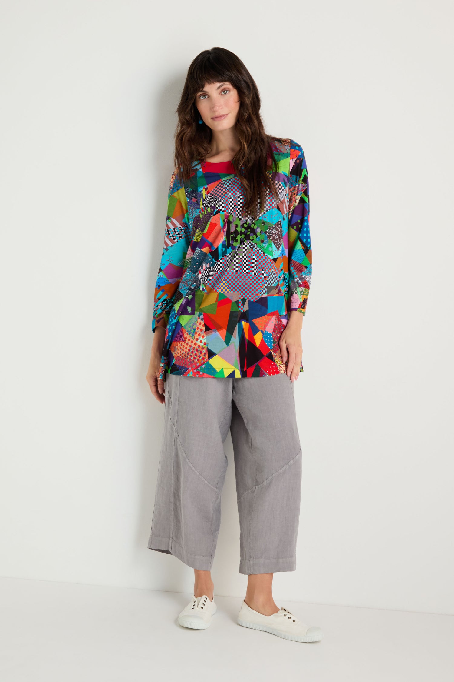 A woman stands against a white wall wearing the Quilt Print Flared Top, featuring multicolored geometric patterns in stretch jersey, paired with gray wide-leg pants and white sneakers.