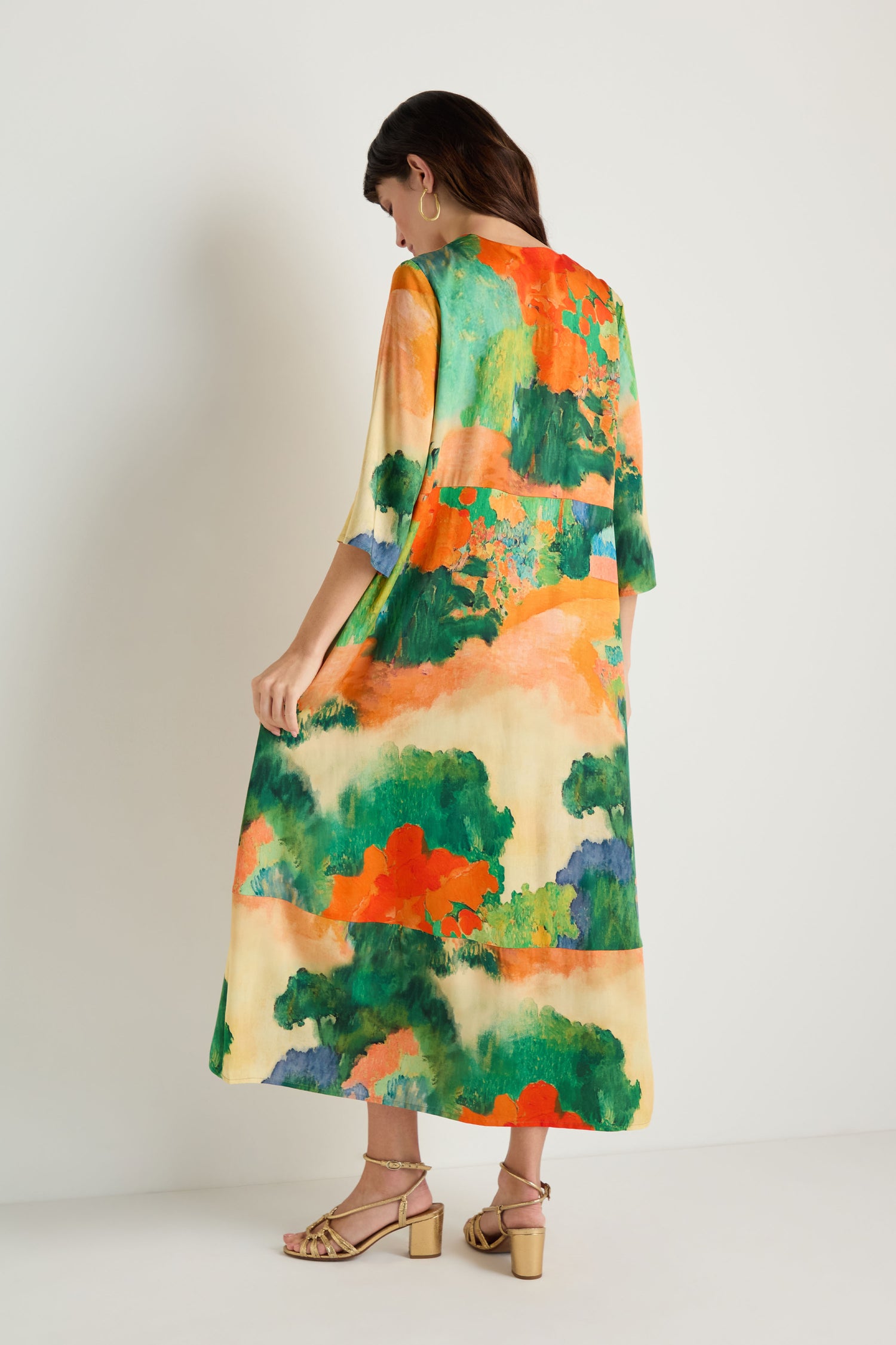A woman stands against a plain wall, wearing the Japanese Garden Flared Dress—long, loose-fitting viscose with an abstract green, orange, and cream pattern—styled with gold heeled sandals.