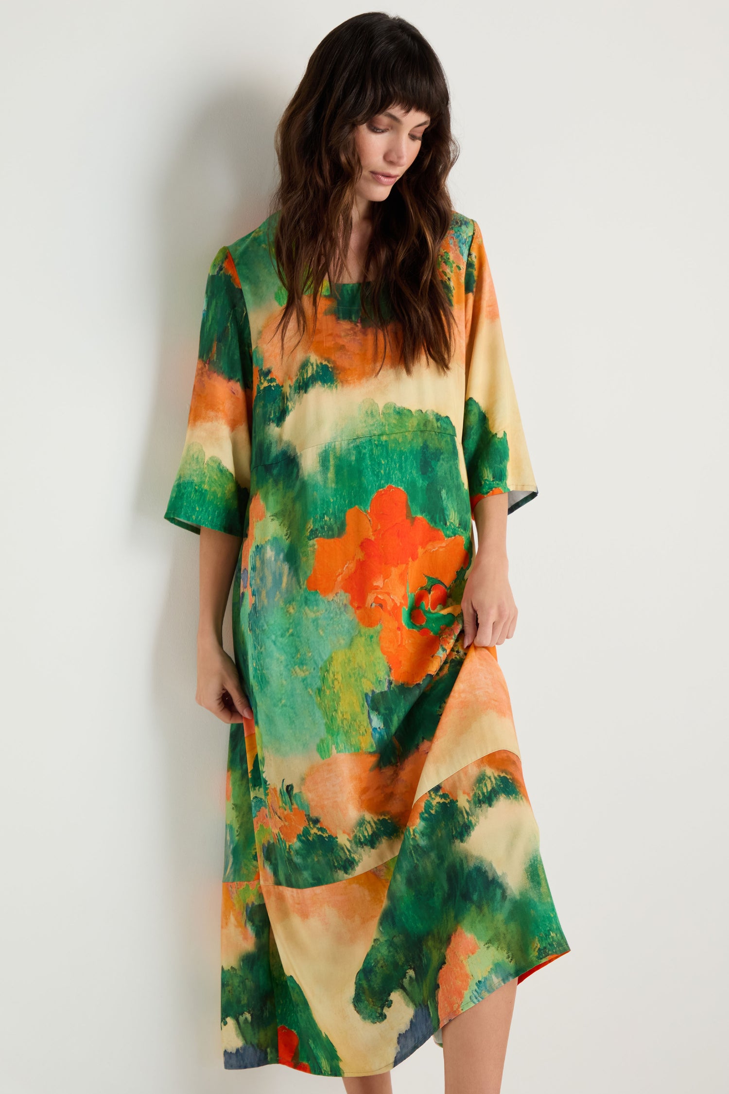 A woman stands against a plain white wall wearing the Japanese Garden Flared Dress, a loose, knee-length piece in woven viscose with colorful abstract green, orange, and yellow patterns.