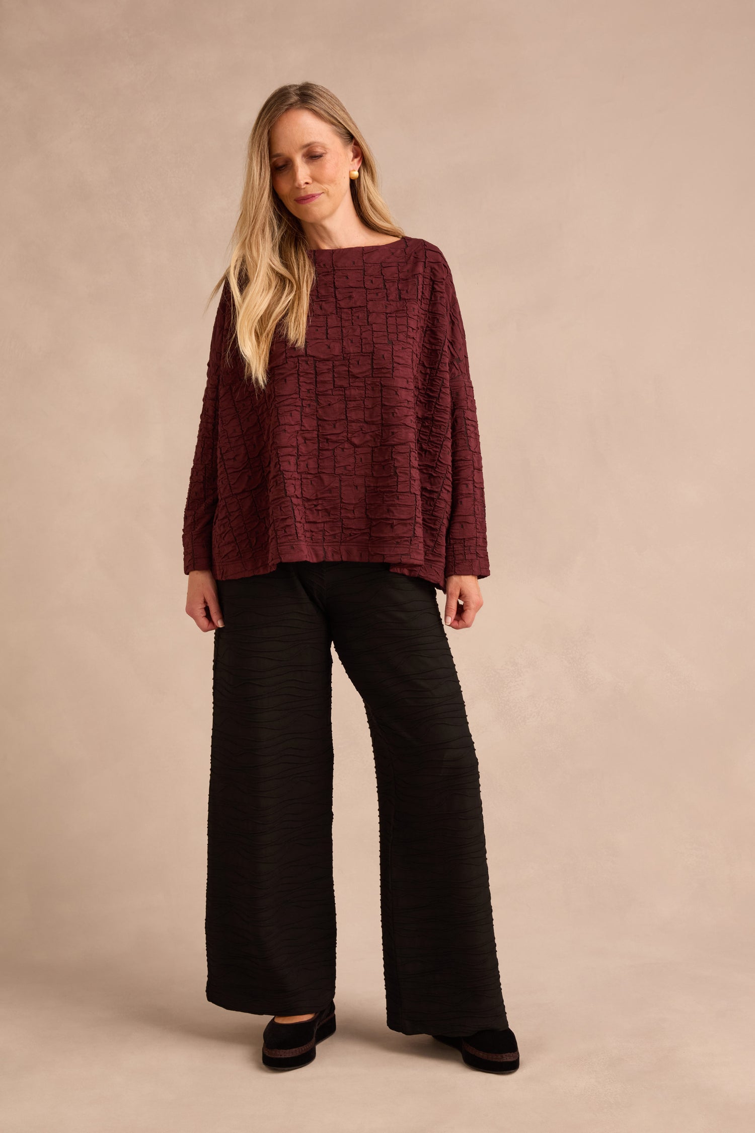 A woman wearing the Bordeaux Textured Squares Jersey Boxy Top and loose black pants stands in front of a neutral beige background.