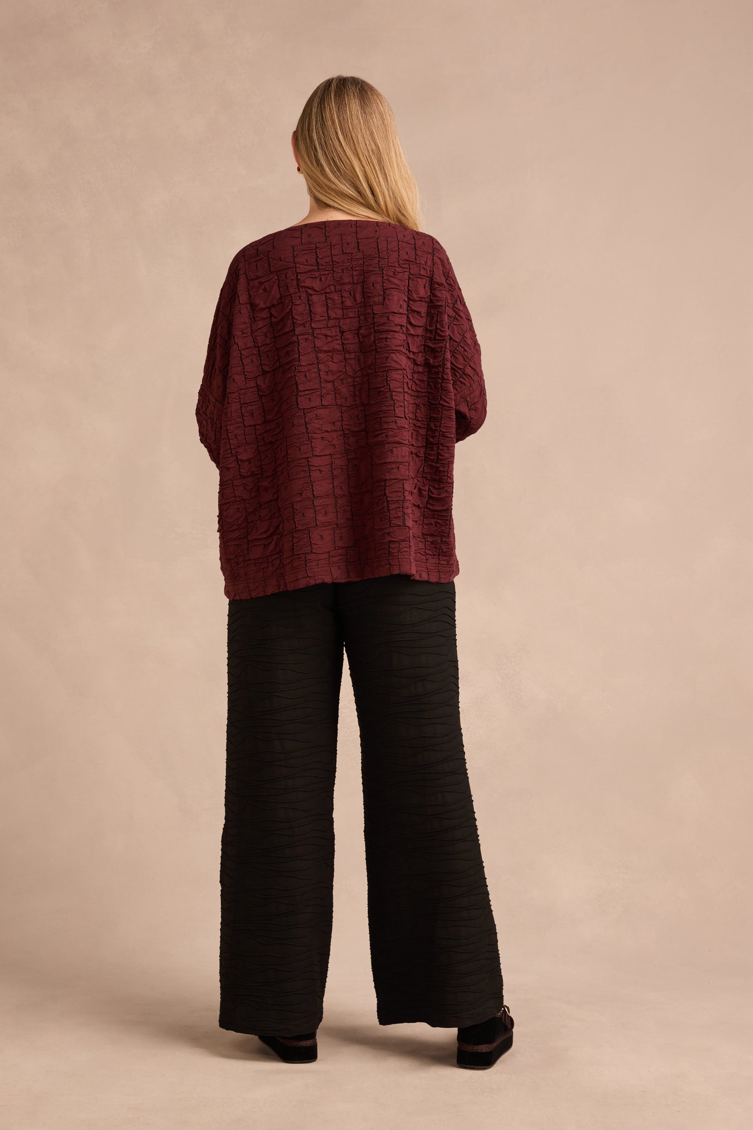 A person stands facing away, wearing the Bordeaux Textured Squares Jersey Boxy Top and loose black pants against a plain beige background.