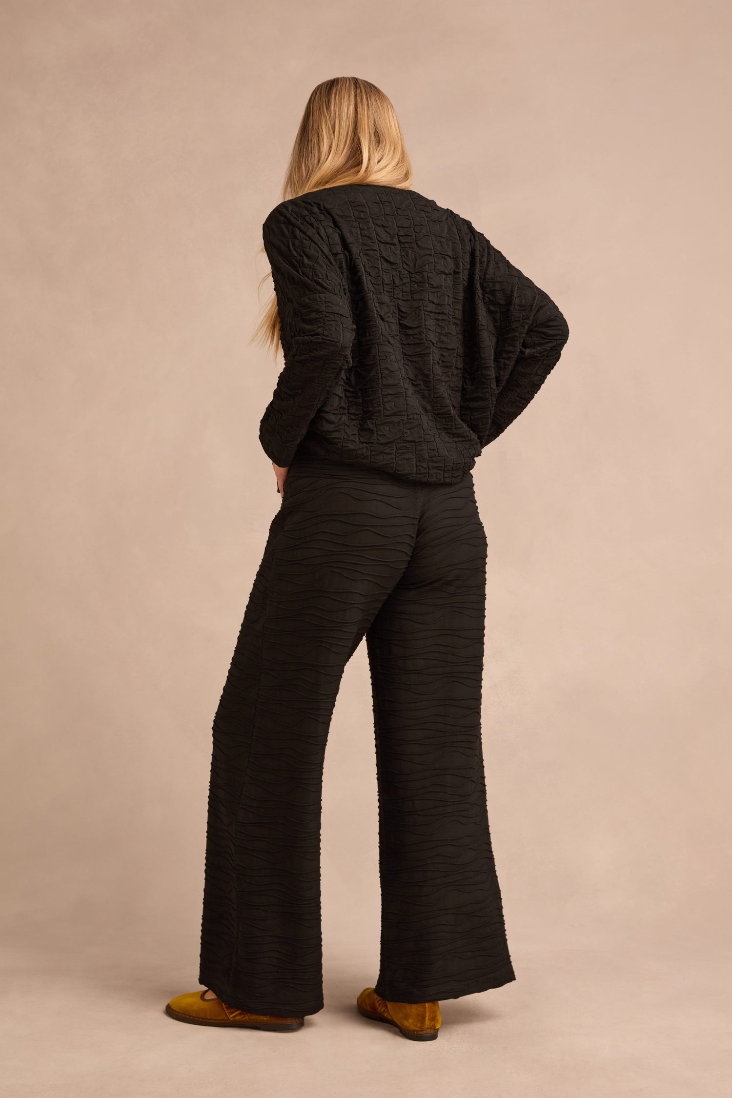 A person with long blond hair stands facing away, wearing the Black Textured Squares Jersey Boxy Top and wide-leg black pants with mustard yellow shoes against a neutral background.