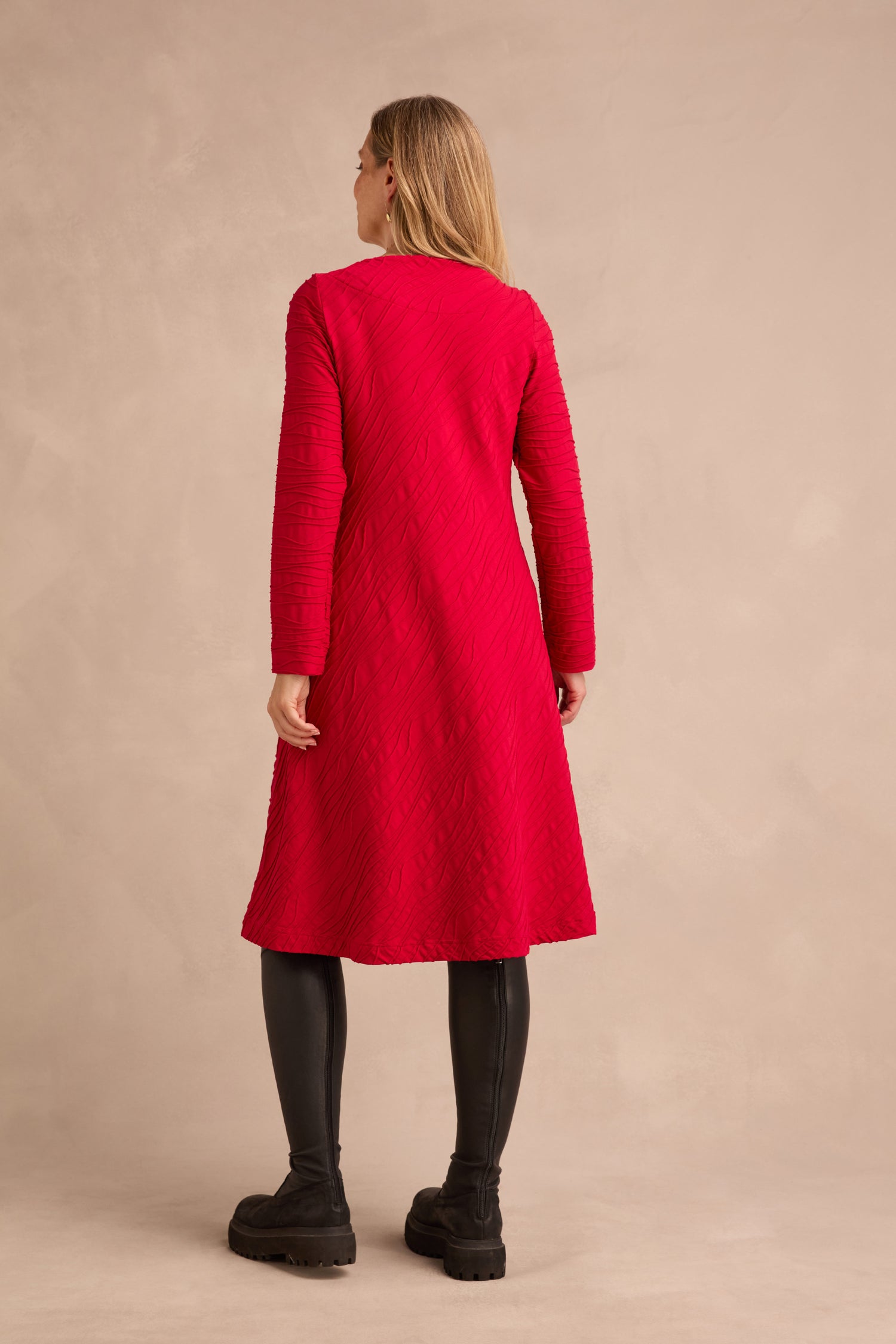 A person stands facing away, wearing the Wave Textured Jersey Flared Dress in a rich red hue, paired with black leggings and chunky black shoes, against a plain beige background.