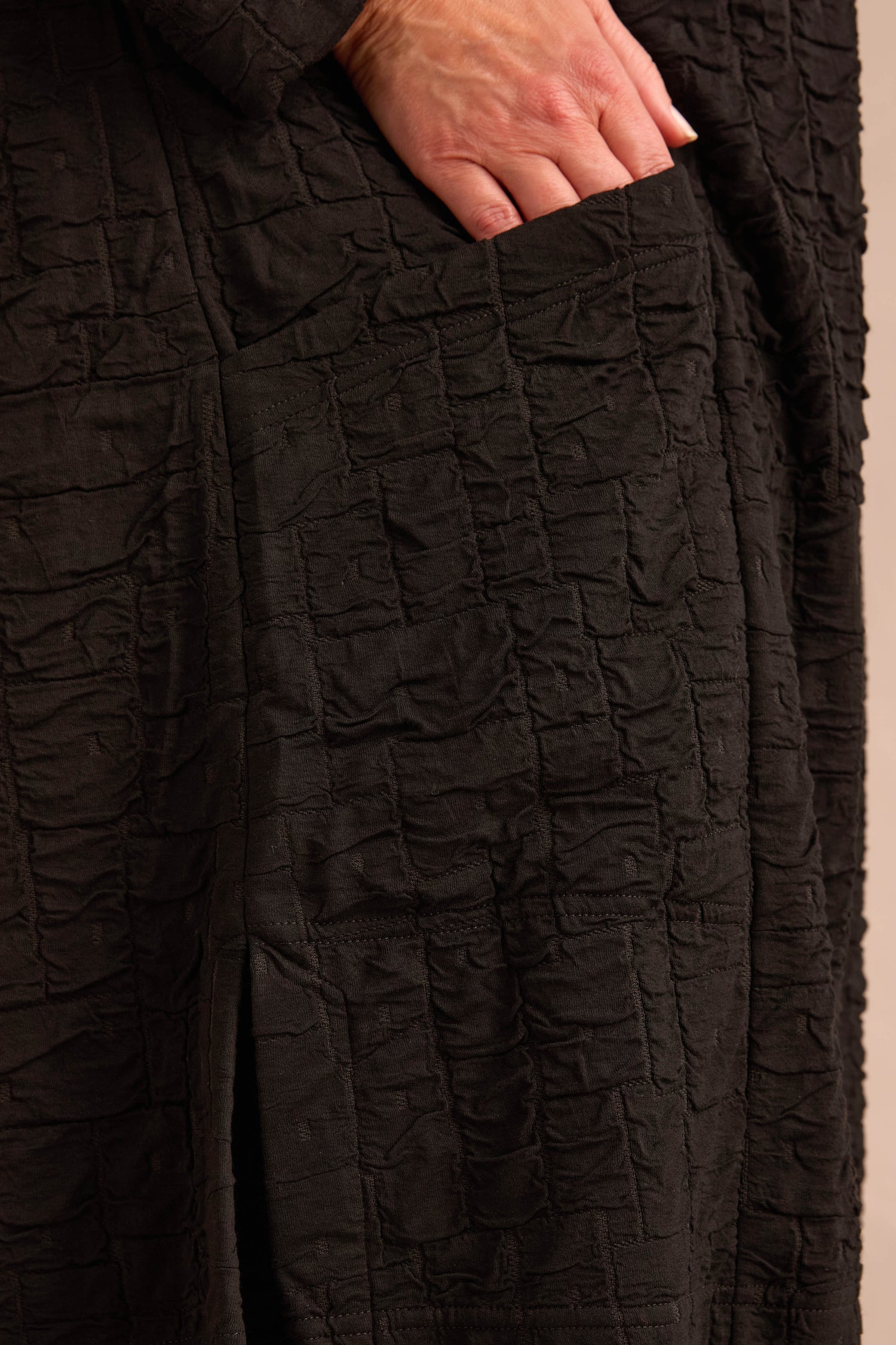 A hand is partially placed inside the pocket of a Textured Squares Jersey Tunic, highlighting the garment’s crinkled pattern in close-up detail.