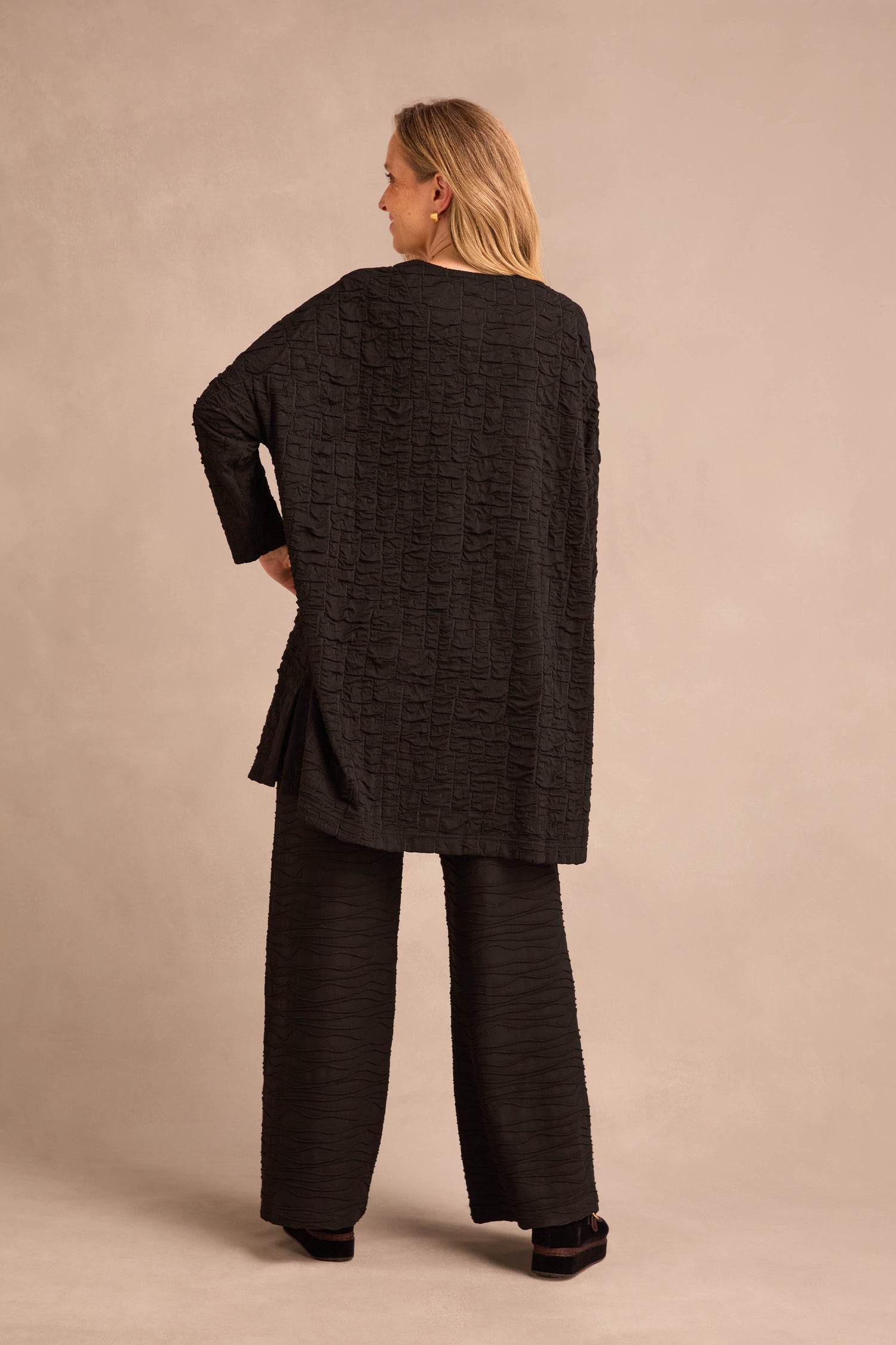 A person stands with their back to the camera, wearing the Textured Squares Jersey Tunic and matching wide-leg pants featuring a subtle square pattern, against a neutral background.