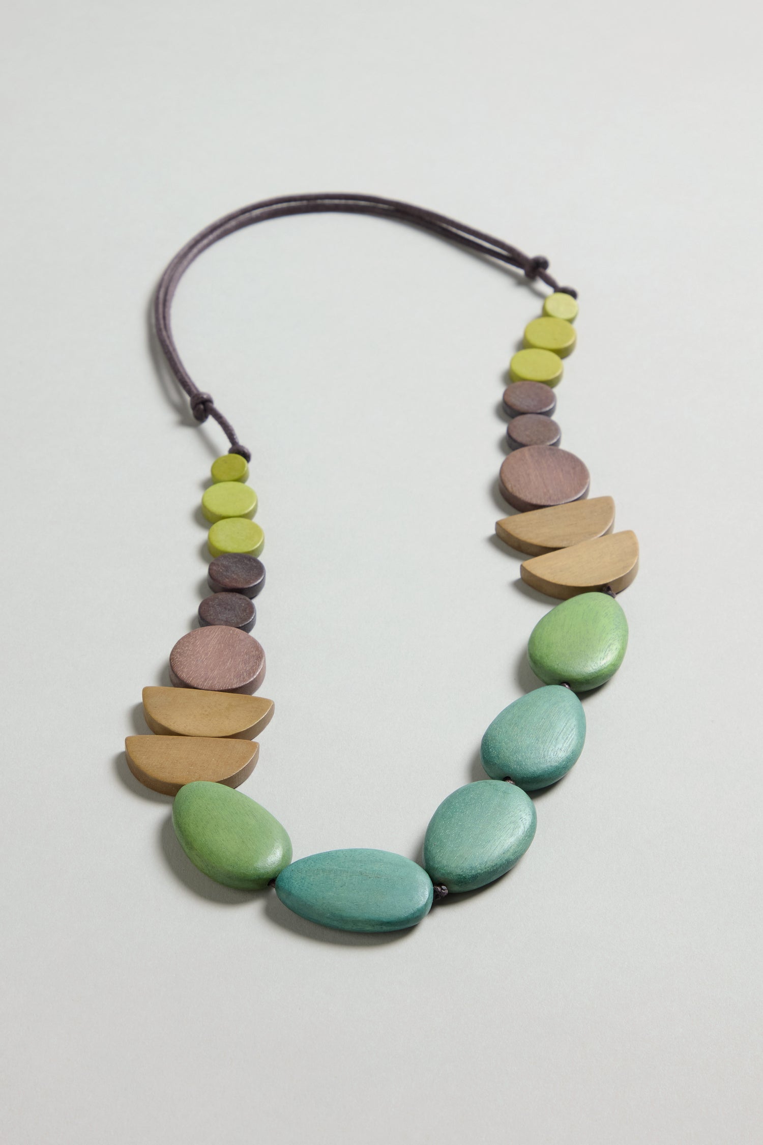 The Assorted Wooden Shapes Necklace features green, beige, brown, and purple wooden beads in various shapes on a dark adjustable cord, displayed on a light gray surface—perfect for making a bold statement.