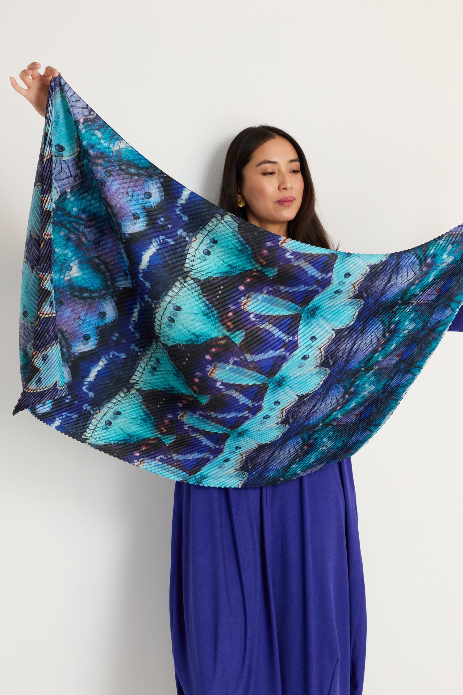 A woman displays the Butterfly Pleat Scarf, a large pleated satin accessory with a blue and purple butterfly wing design, standing out as a lightweight statement piece against a plain white background.