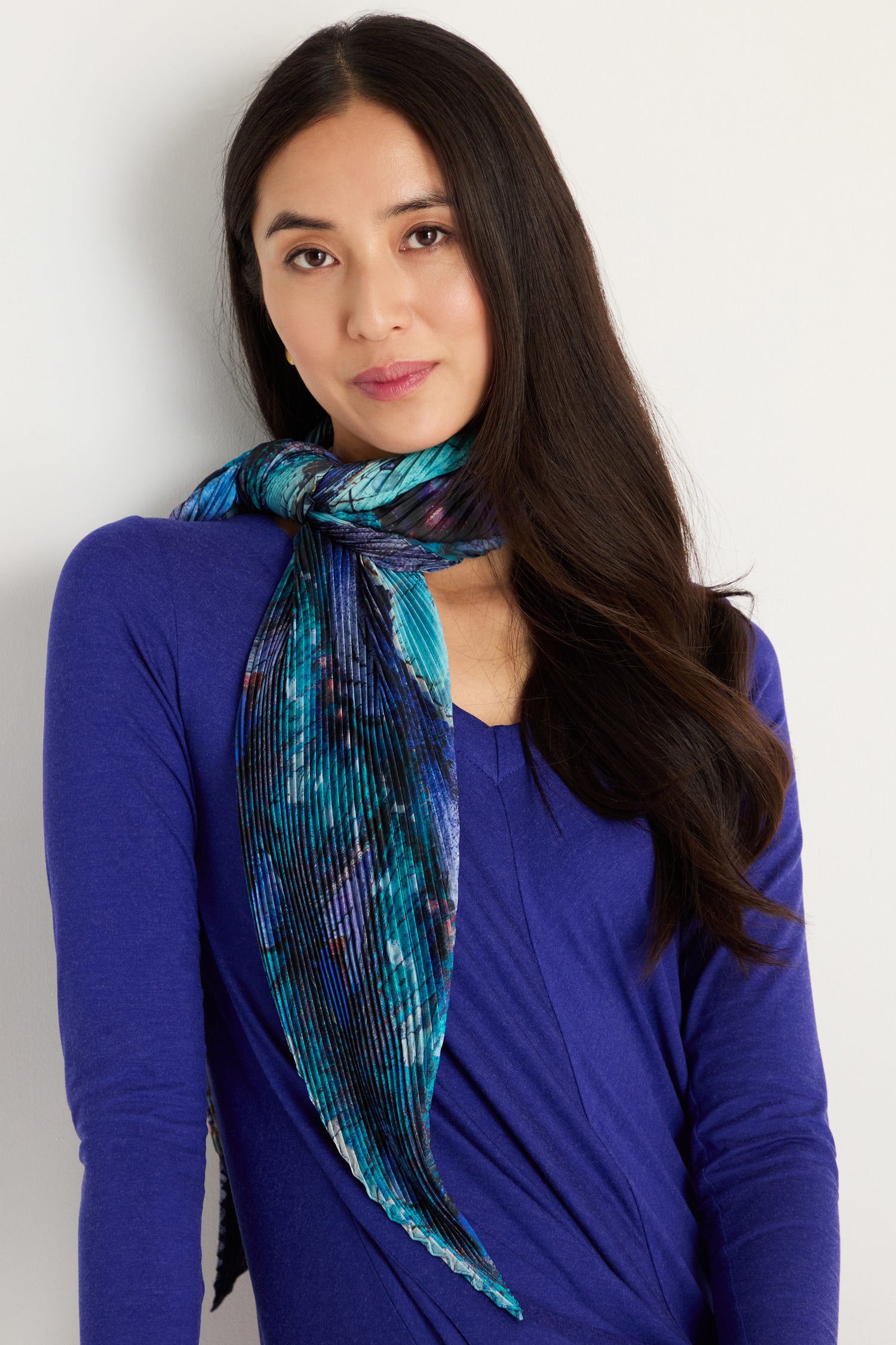 A woman with long dark hair, wearing a blue top and the Butterfly Pleat Scarf tied around her neck, stands against a plain light background.
