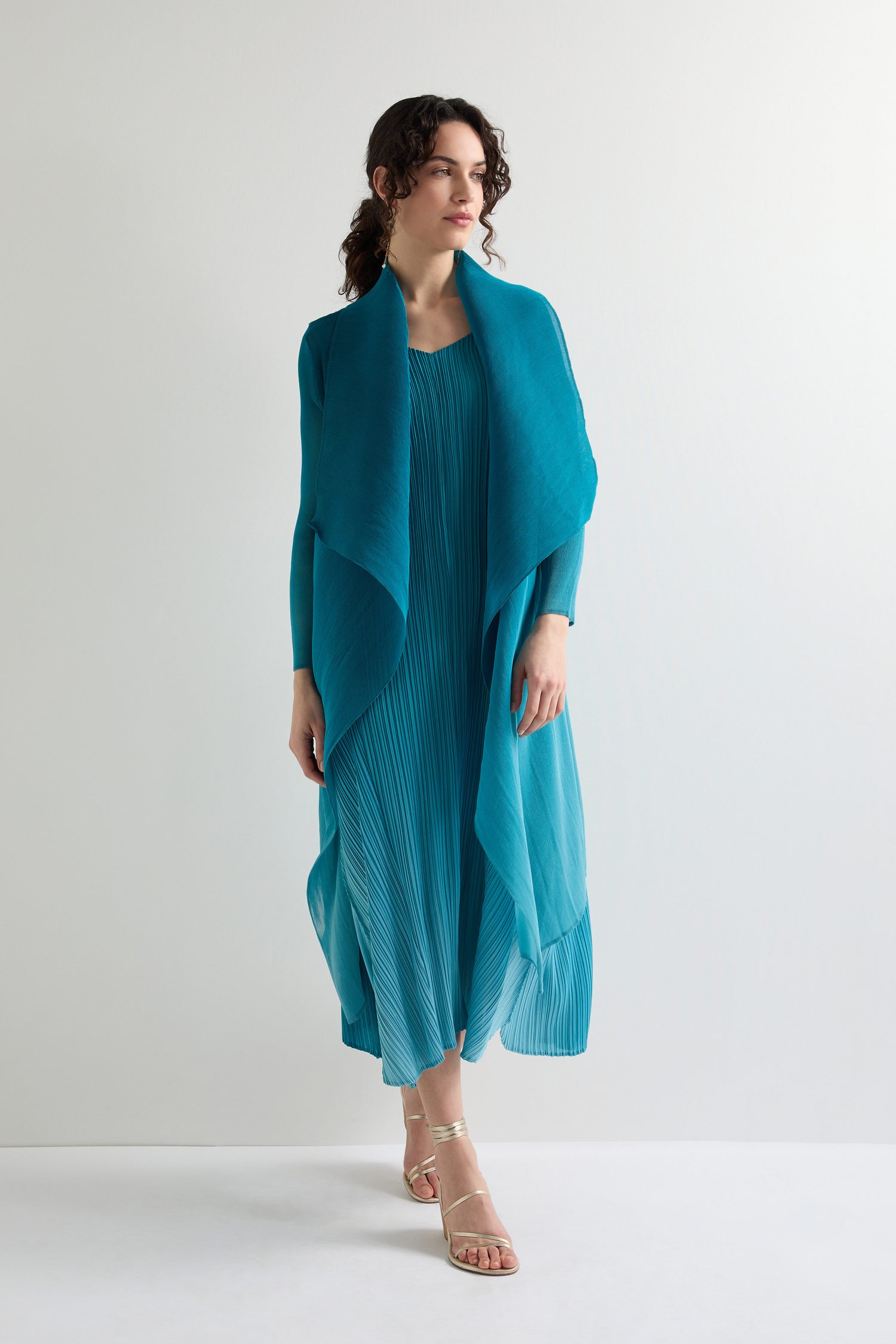 A person in a flowing teal dress and Pleat Jacket stands against a plain backdrop, exuding bohemian flair with their choice of sandals.