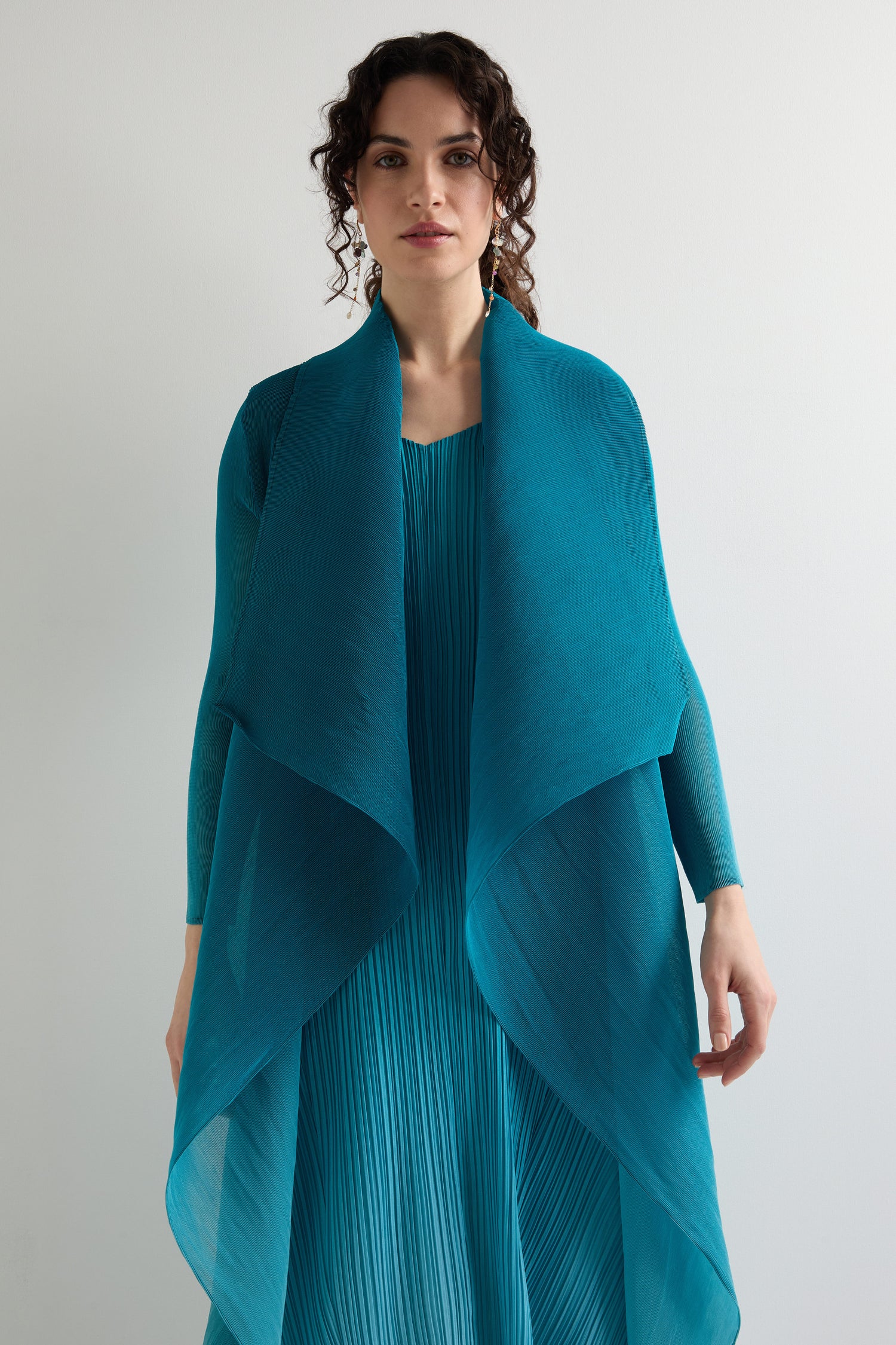 A person in a teal, pleated "Pleat Jacket" is set against a plain white background, giving off bohemian vibes.