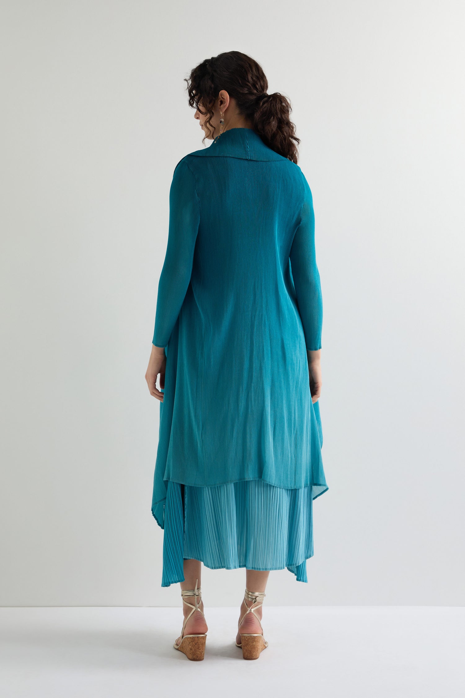 A person radiates modern style in a teal Pleat Jacket, long-sleeved and standing with their back to a plain white wall.