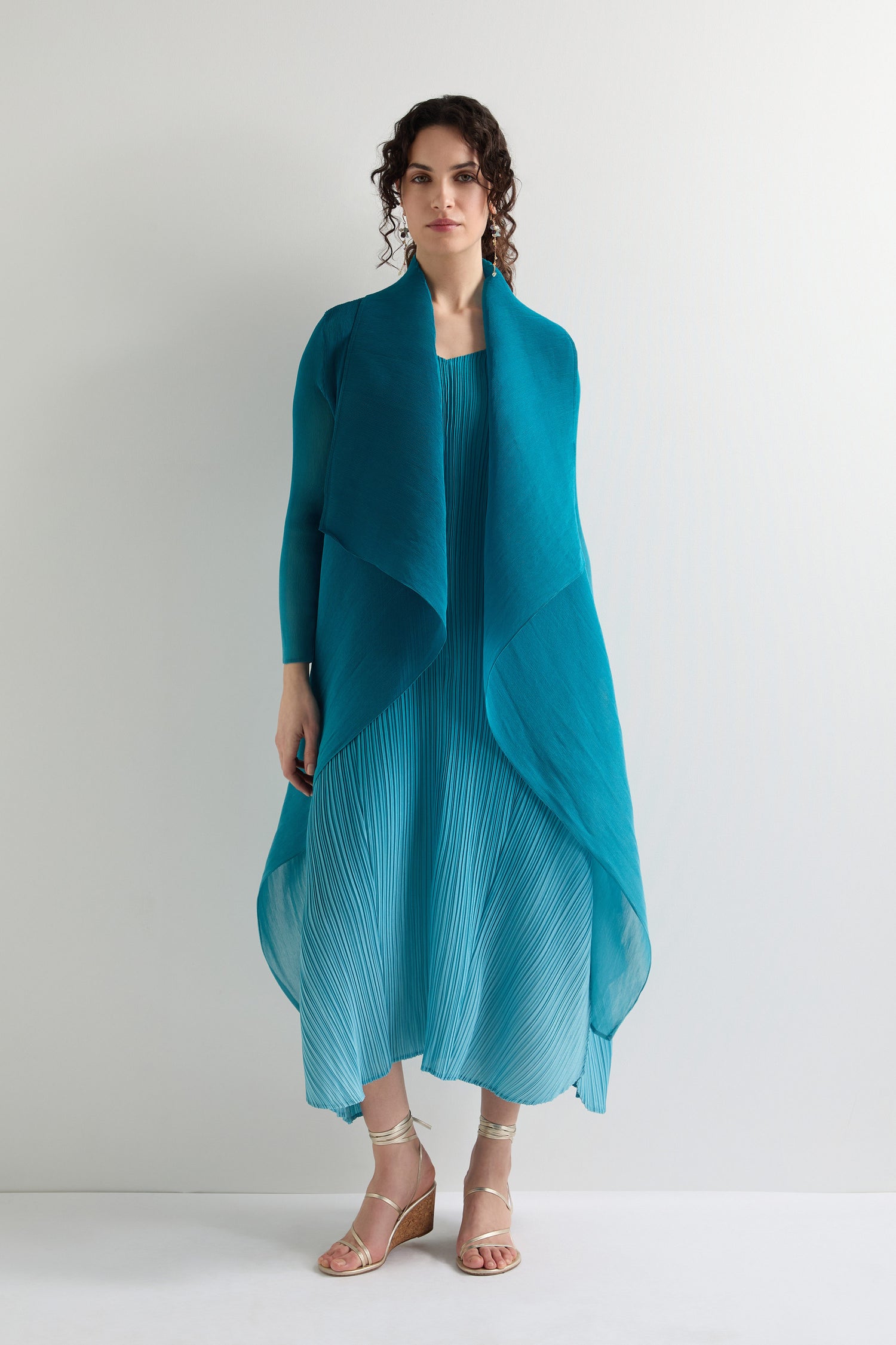 Wearing a teal pleated dress with a matching Pleat Jacket, the person exudes contemporary style against a light background.