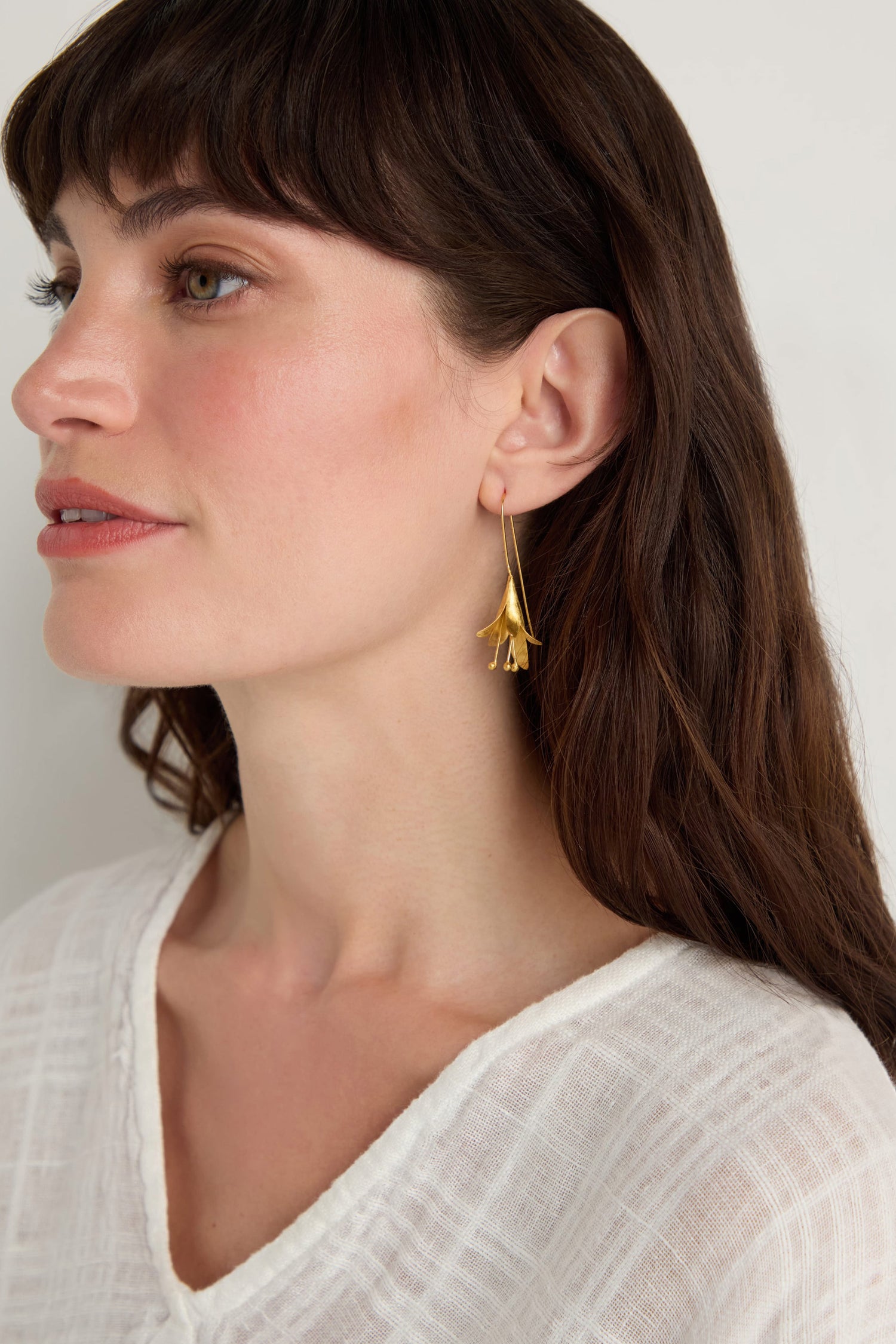 A woman with long brown hair wears a white textured top and the Gold Vermeil Flower Earrings, delicately shaped like flowers, as she faces left against a neutral background.