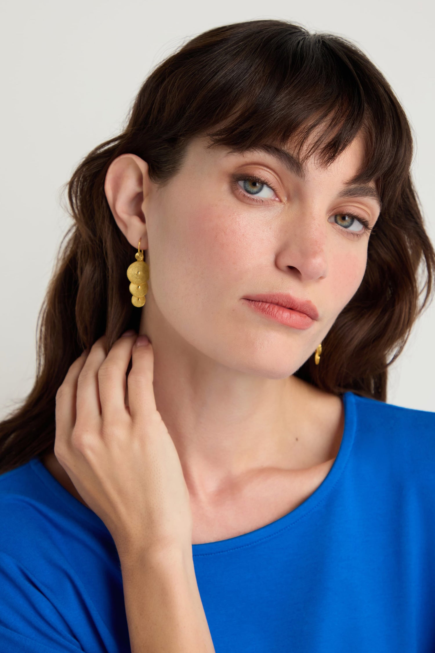 Woman with long brown hair and bangs, wearing a blue shirt and Gold Vermeil Orbit Disc Earrings, looks at the camera with a neutral expression while touching her neck.