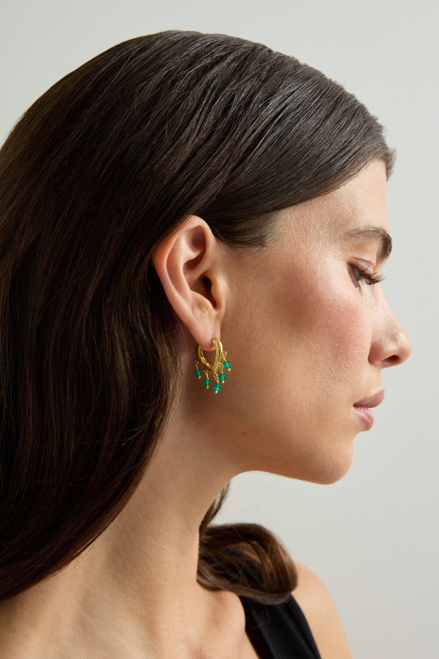 A woman with straight brown hair is shown in profile against a plain light background, wearing Gold Vermeil Onyx Teardrop Hoops featuring green onyx teardrop pendants.