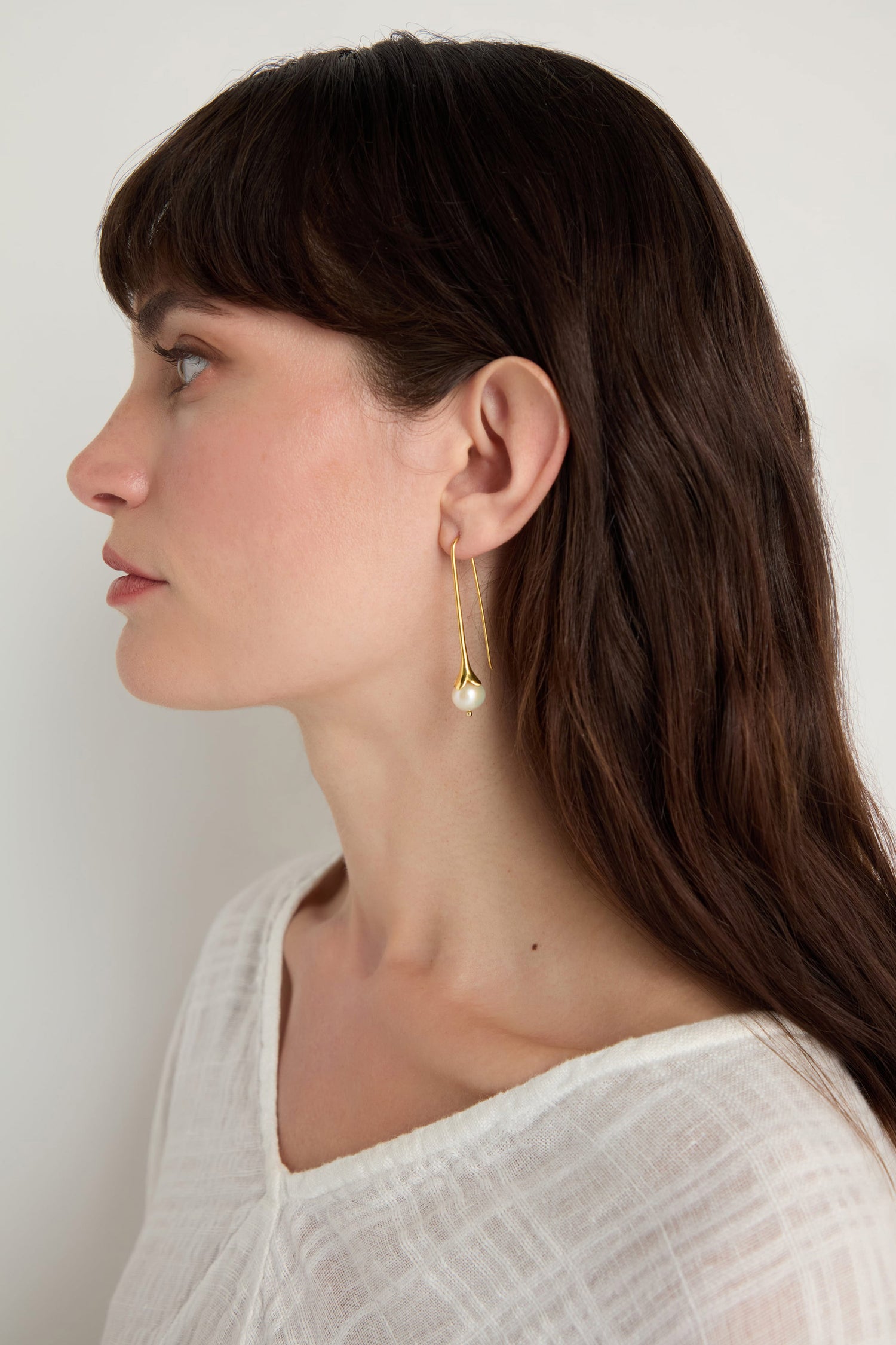 Woman with long brown hair, in a white textured top, wears the Gold Vermeil Pearl Drop Earrings and is shown in left profile against a plain background.