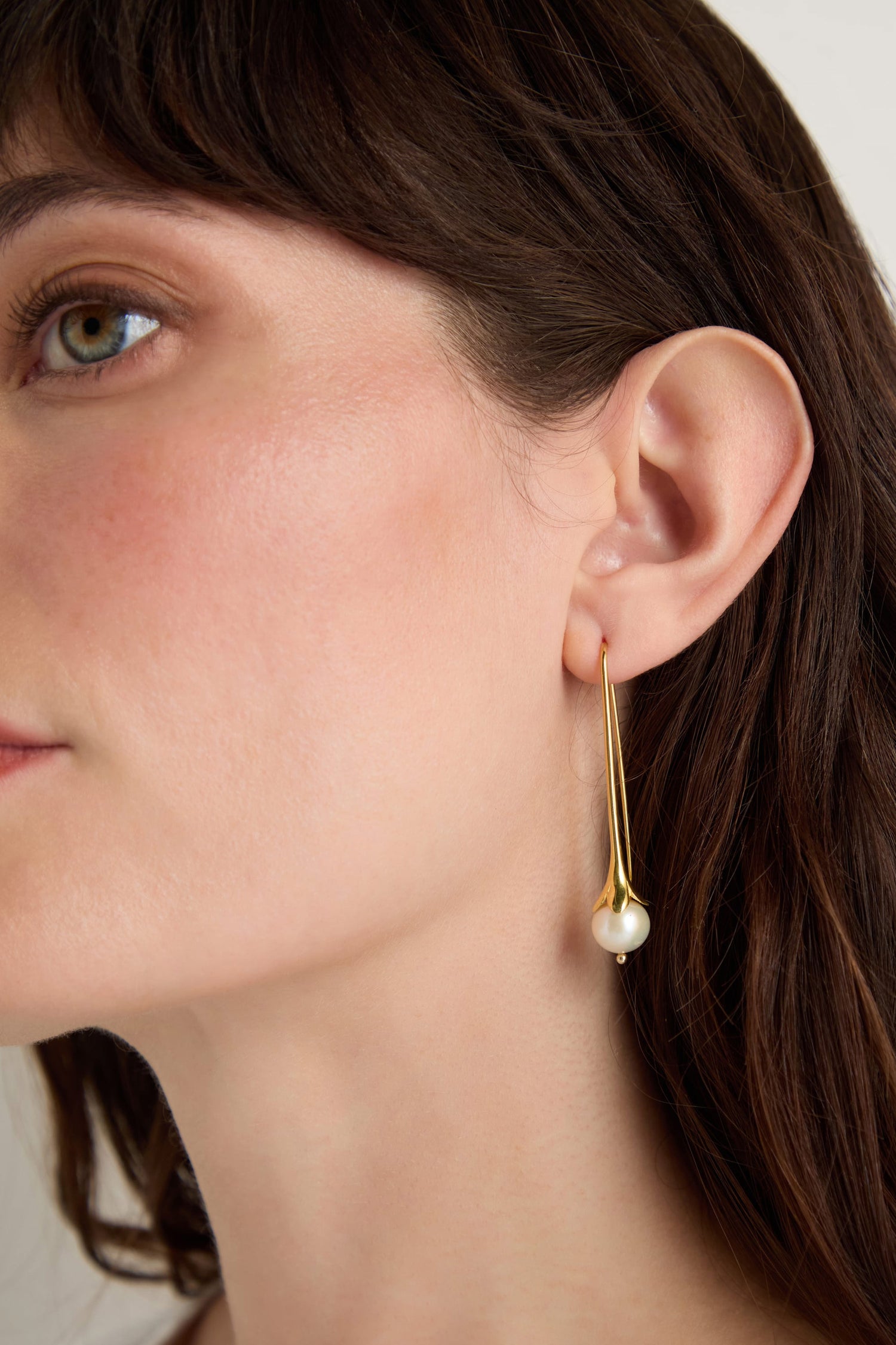 Close-up of a woman wearing Gold Vermeil Pearl Drop Earrings on her left ear, featuring a dangling pearl, set against a neutral background.