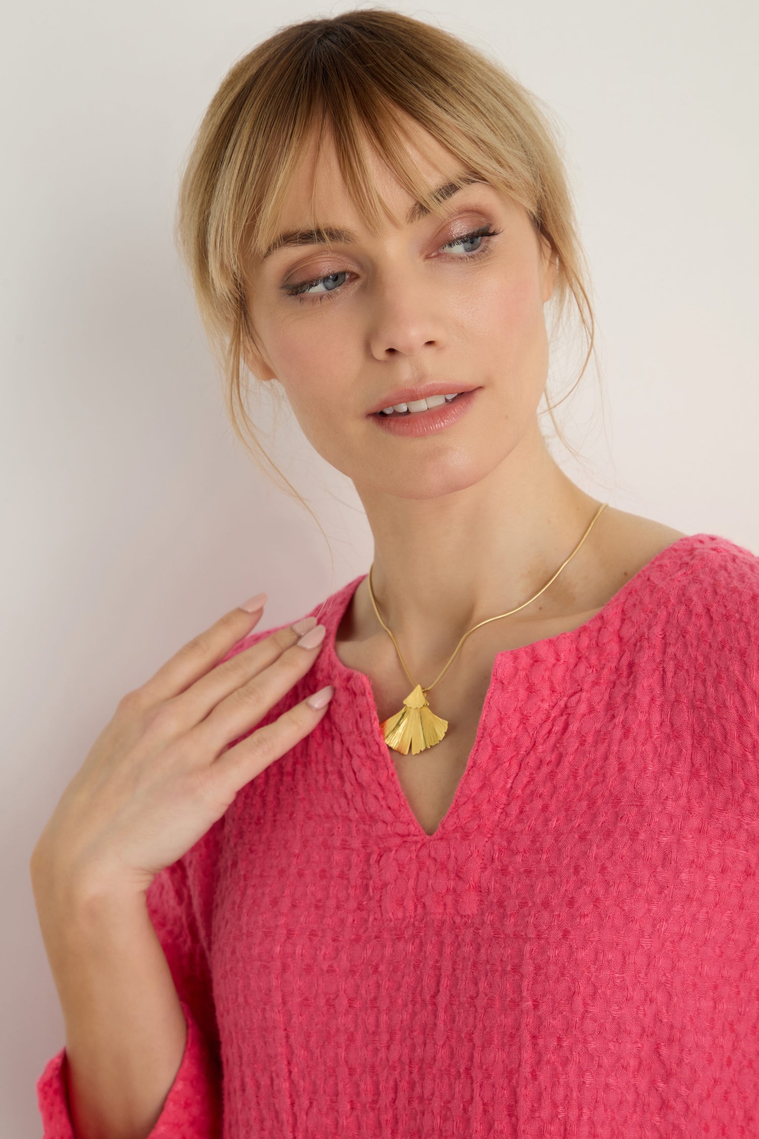 A woman in a textured pink top wears the Gold Vermeil Ginkgo Leaf Pendant necklace, gazing slightly to the side with one hand raised near her neck.