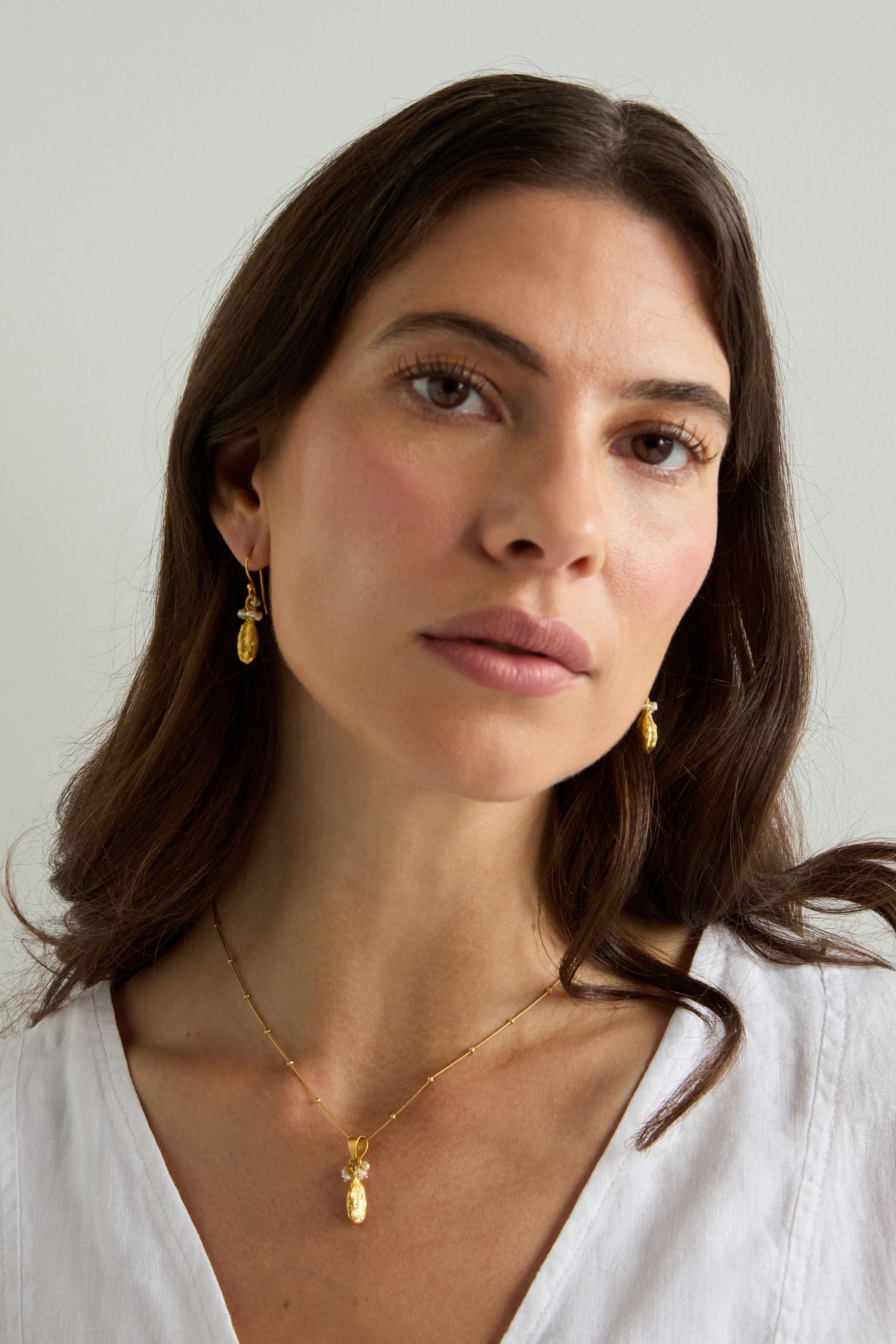 A woman with long brown hair wears Gold Vermeil Pearl Drop Earrings and a gold necklace, paired with a white top against a plain light background.
