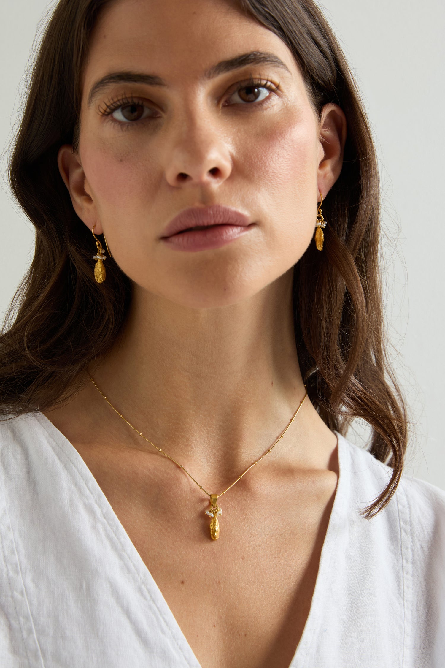 A woman with long brown hair wears a white V-neck blouse, gold drop earrings, and the Gold Vermeil Pearl Drop Necklace, looking directly at the camera against a plain background.