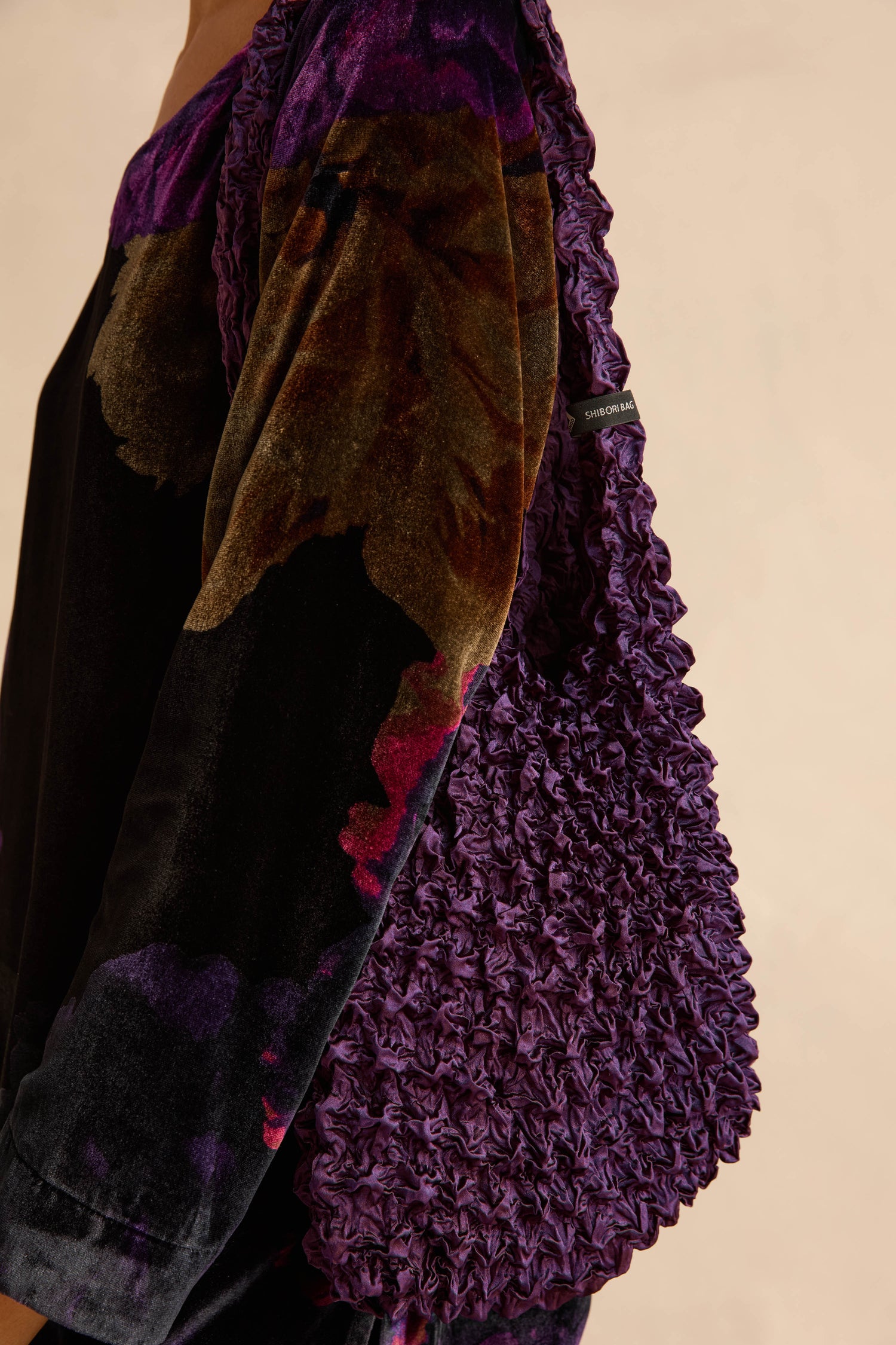 A person wears a dark, patterned velvet garment while carrying an Aubergine Shibori Pleated Bag, a deep purple, pleated accessory with a heavily gathered, textured surface.